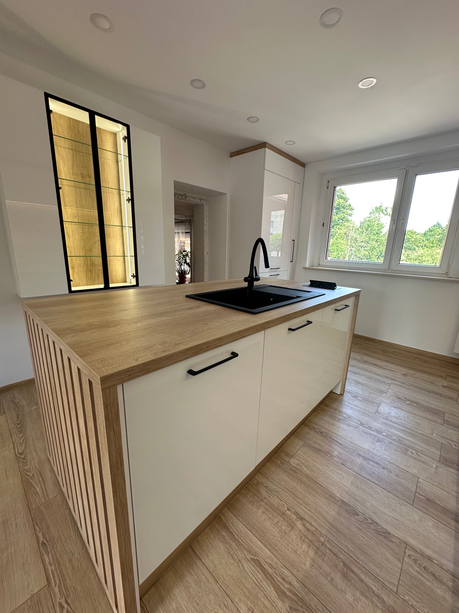 Nowoczesna wyspa kuchenna z blatem imitującym drewno, czarnym zlewem i minimalistycznymi uchwytami, na tle białych szafek i okna z widokiem na zieleń.