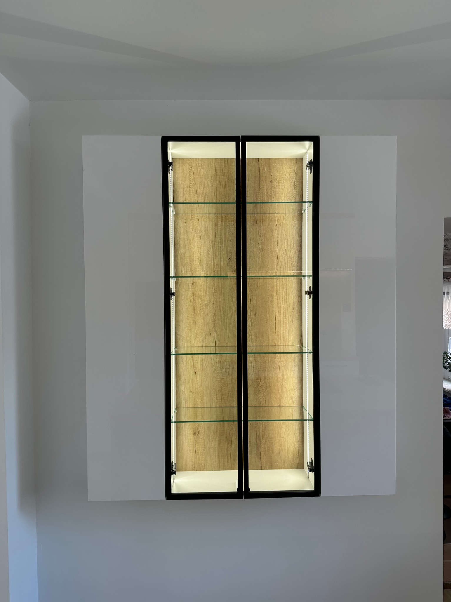 Wisząca szafka z podświetleniem LED i drewnianym wnętrzem, wbudowana w białą ścianę. Minimalistyczny design, szklane półki, czarne obramowanie drzwi.