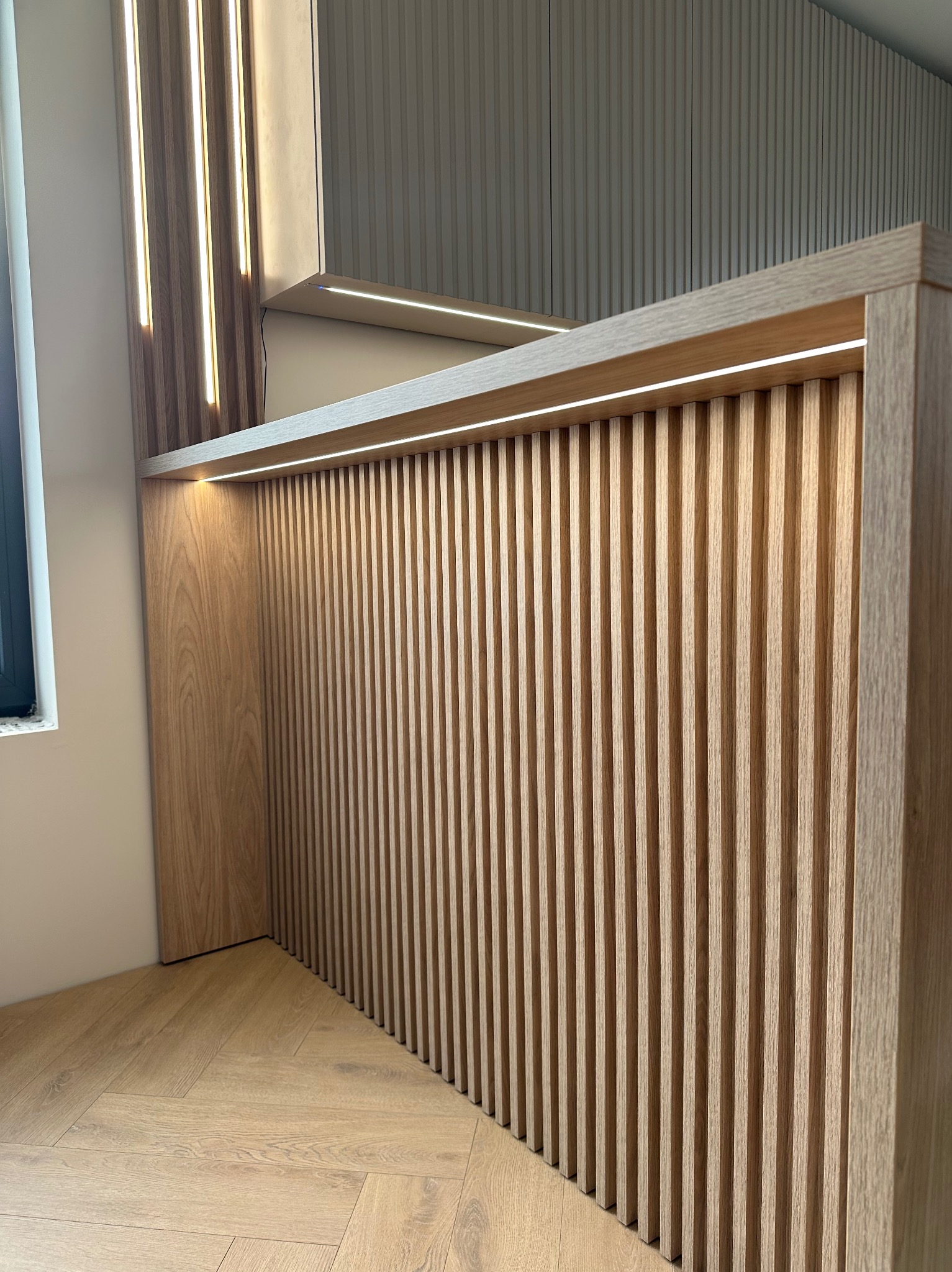 Nowoczesna zabudowa meblowa z ryflowanym frontem i oświetleniem LED, wykonana z drewna w jasnym odcieniu dębu, z widocznym fragmentem podłogi w jodełkę.