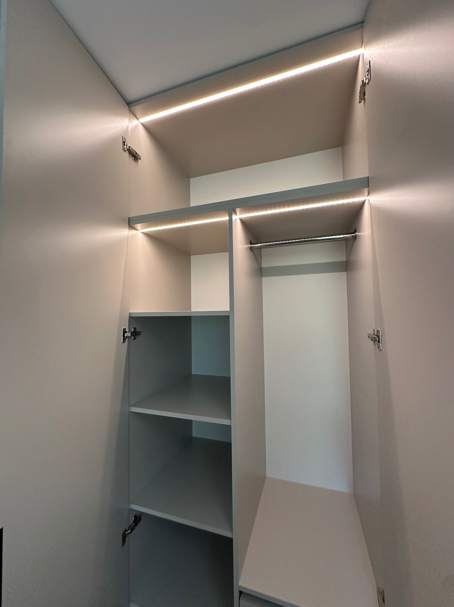 Otwarta szafa wnękowa z półkami i drążkiem, oświetlona listwami LED. Widoczne zawiasy i jasne wnętrze szafy. Ujęcie od dołu, wnętrze szafy w odcieniach szarości.