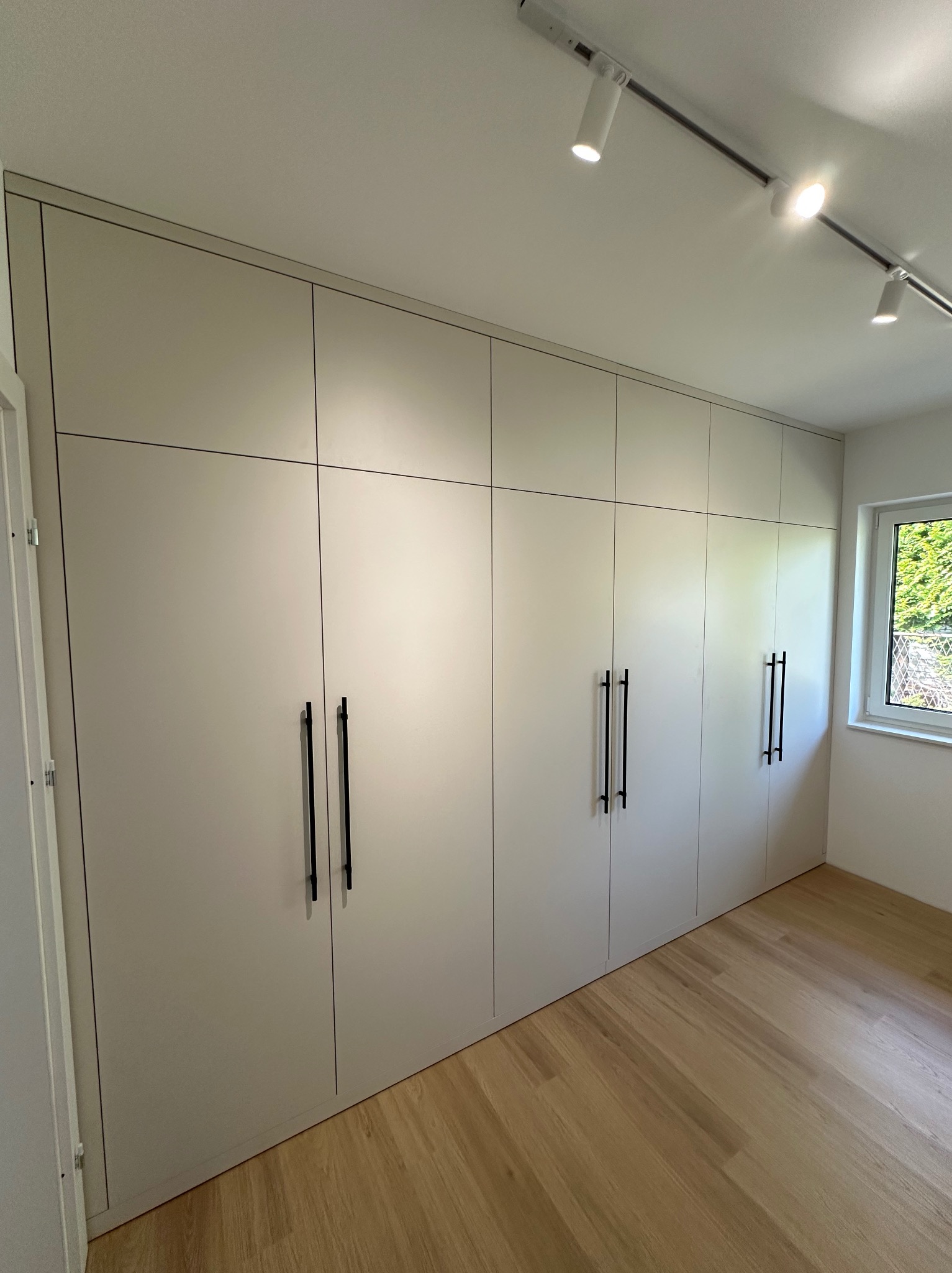 Jasna szafa wnękowa z czarnymi, minimalistycznymi uchwytami, wkomponowana w przestrzeń pokoju z drewnianą podłogą i oświetleniem punktowym.