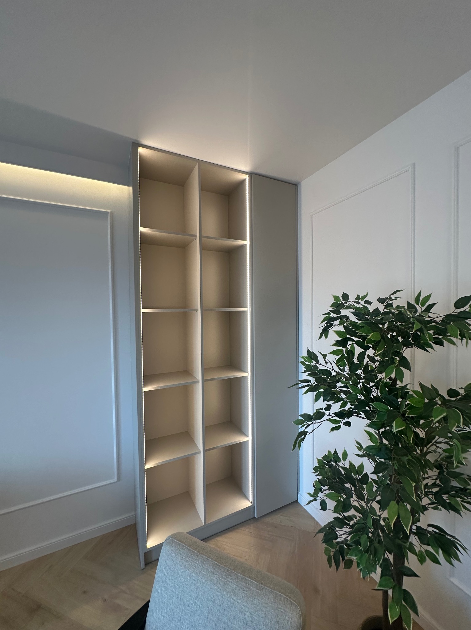 Wysoka, beżowa szafa wnękowa z oświetleniem LED, wkomponowana w ścianę z dekoracyjnymi listwami. Obok stoi doniczkowa roślina, a w kadrze widoczny fragment fotela.
