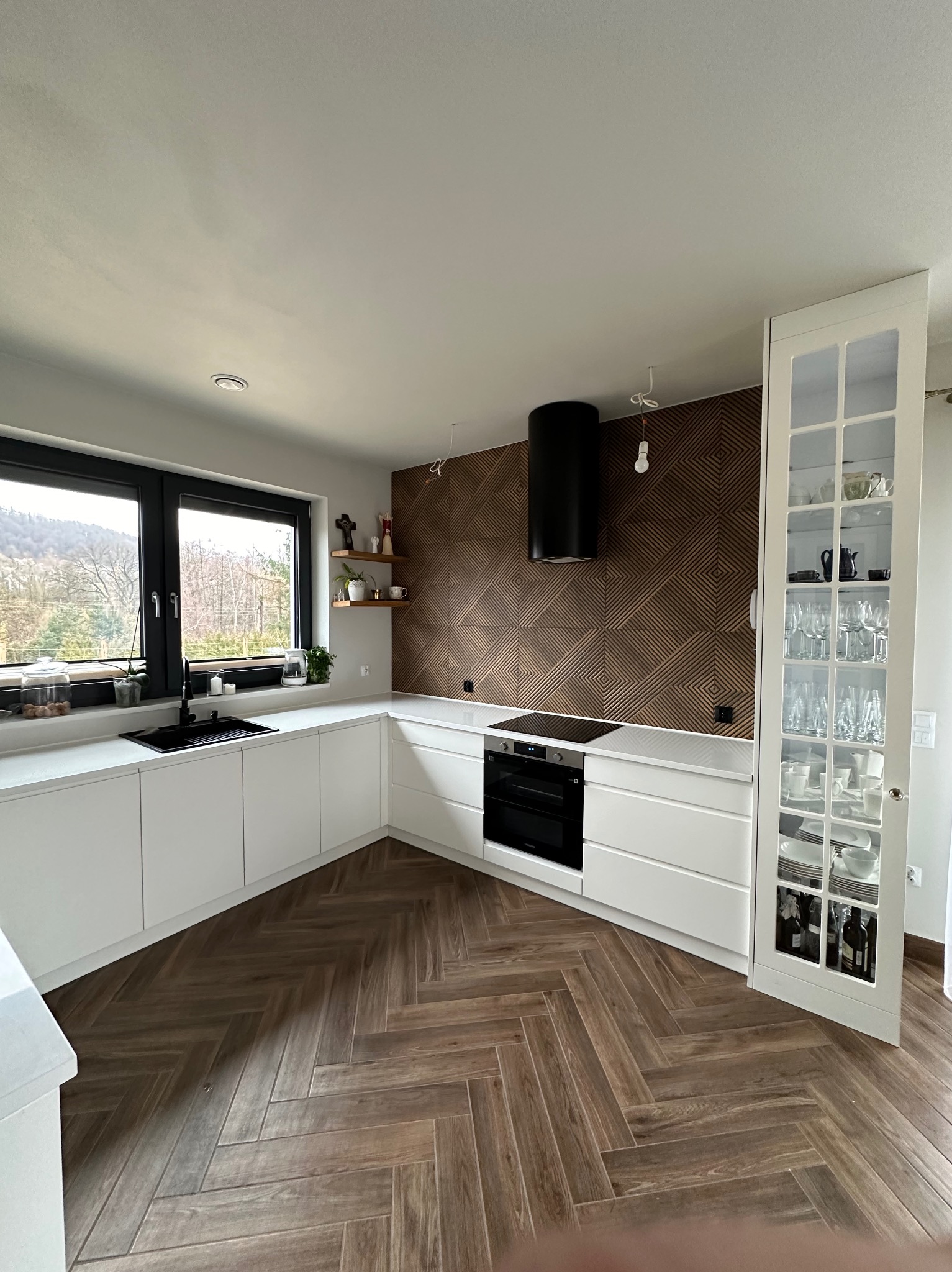 Nowoczesna kuchnia z białymi frontami, drewnianą ścianą dekoracyjną i podłogą w jodełkę. Na blacie przy oknie widoczne rośliny i akcesoria kuchenne.