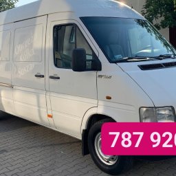 Cokolwiek - Biały van Volkswagen LT35 z napisem 'Obrzeża Kółka Suszarka' na boku, zaparkowany na kostce brukowej, z numerem telefonu nałożonym na dolną część zdjęcia.