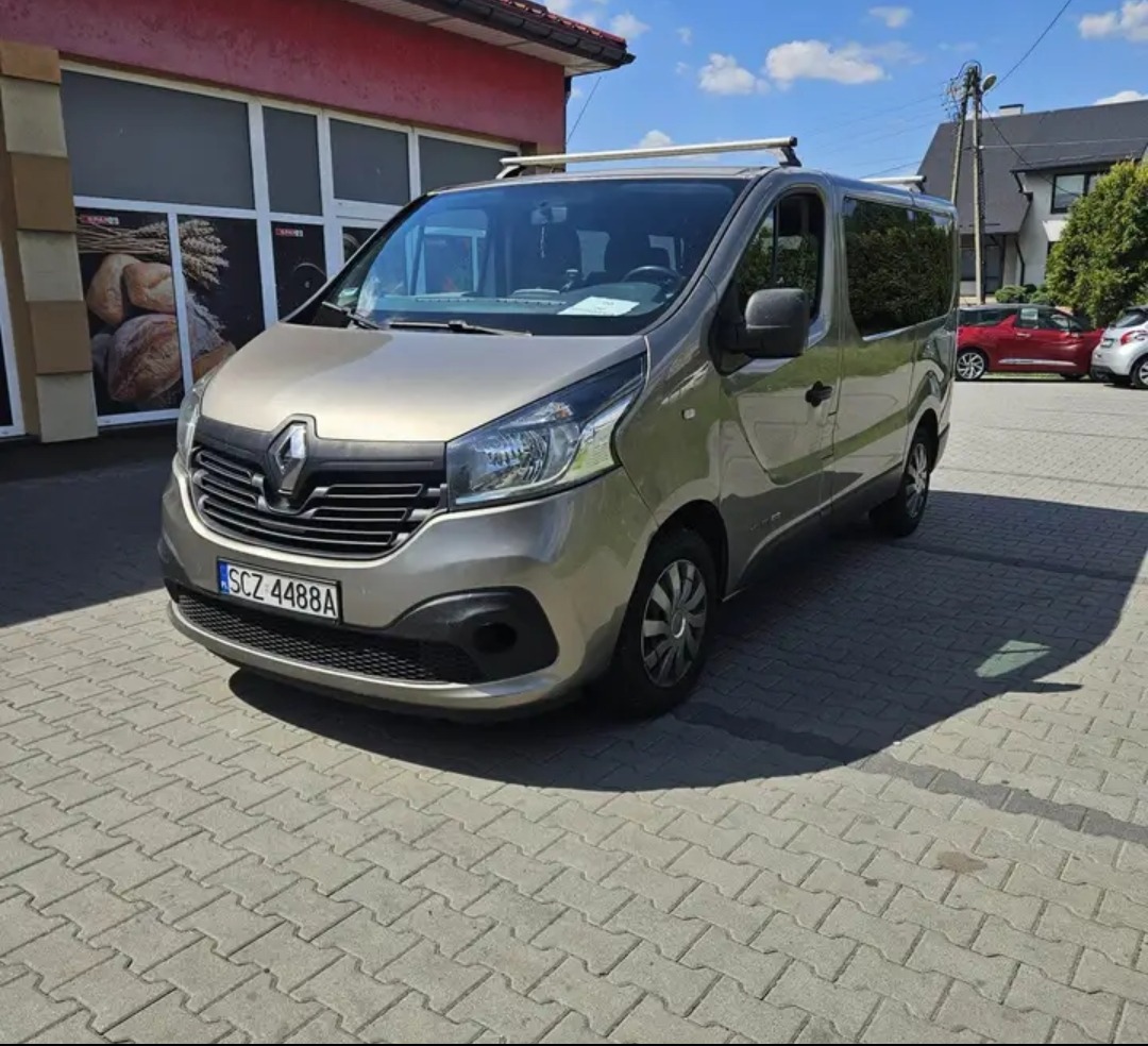 Szary Renault Trafic z bagażnikiem dachowym zaparkowany na kostce brukowej przed budynkiem z oknami. Widoczny fragment tablicy rejestracyjnej.