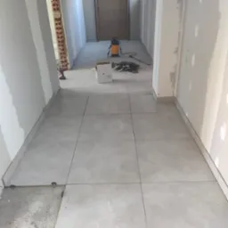 Układanie jasnoszarej, dużej glazury w korytarzu. Widoczne narzędzia i materiały budowlane. Częściowo zakończone prace wykończeniowe ścian.