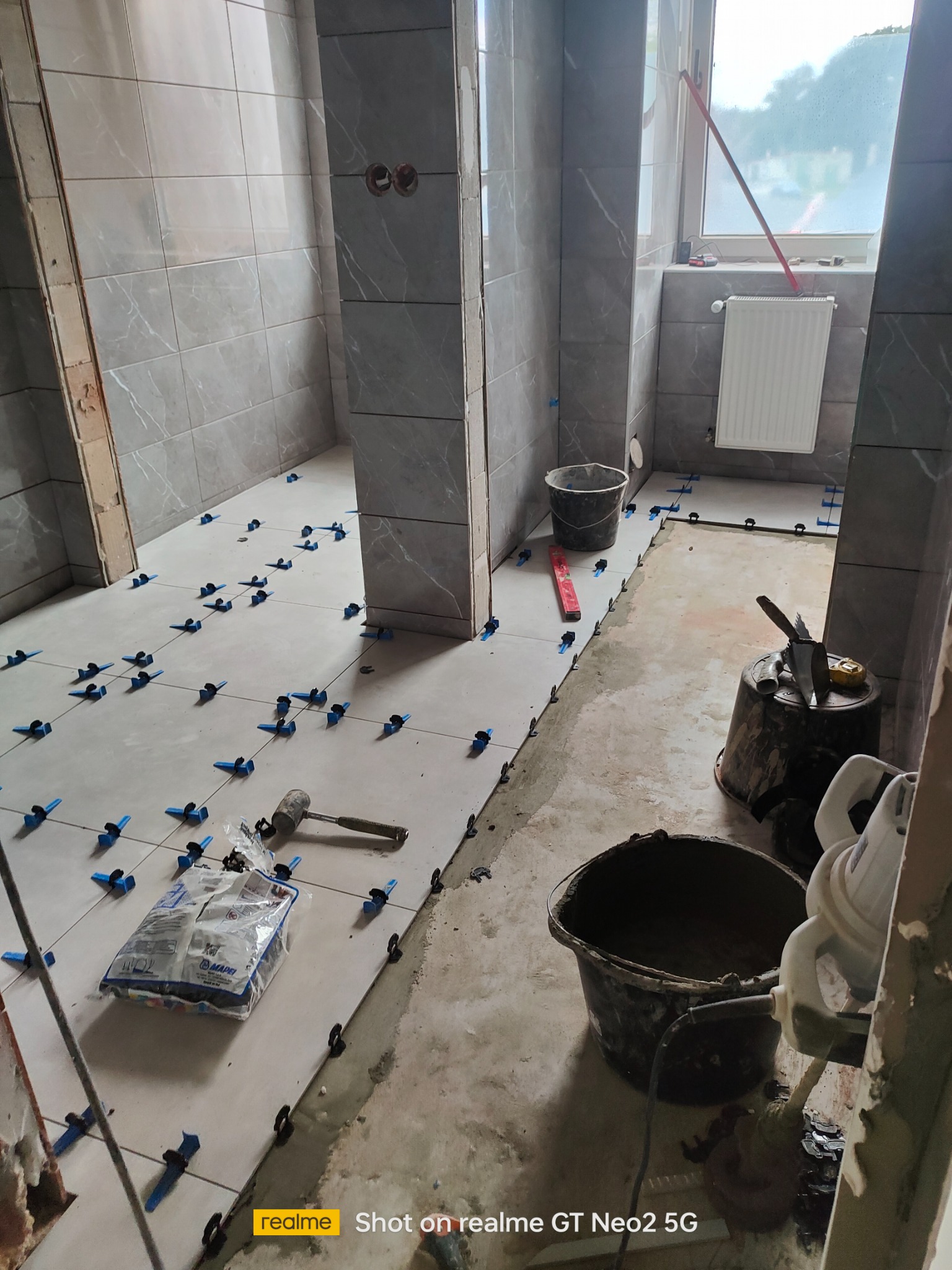 Wnętrze łazienki w trakcie prac glazurniczych: szare płytki na ścianach i podłodze, system poziomowania płytek, narzędzia glazurnicze i wiadro z zaprawą. Częściowo wykończone.