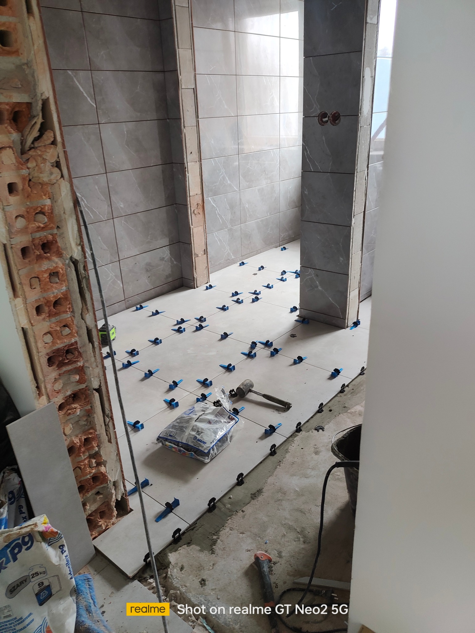 Układanie szarych płytek podłogowych z użyciem niebieskich klipsów poziomujących. Widoczny młotek gumowy, wiadro i worek z klejem. Surowe ściany z cegły.