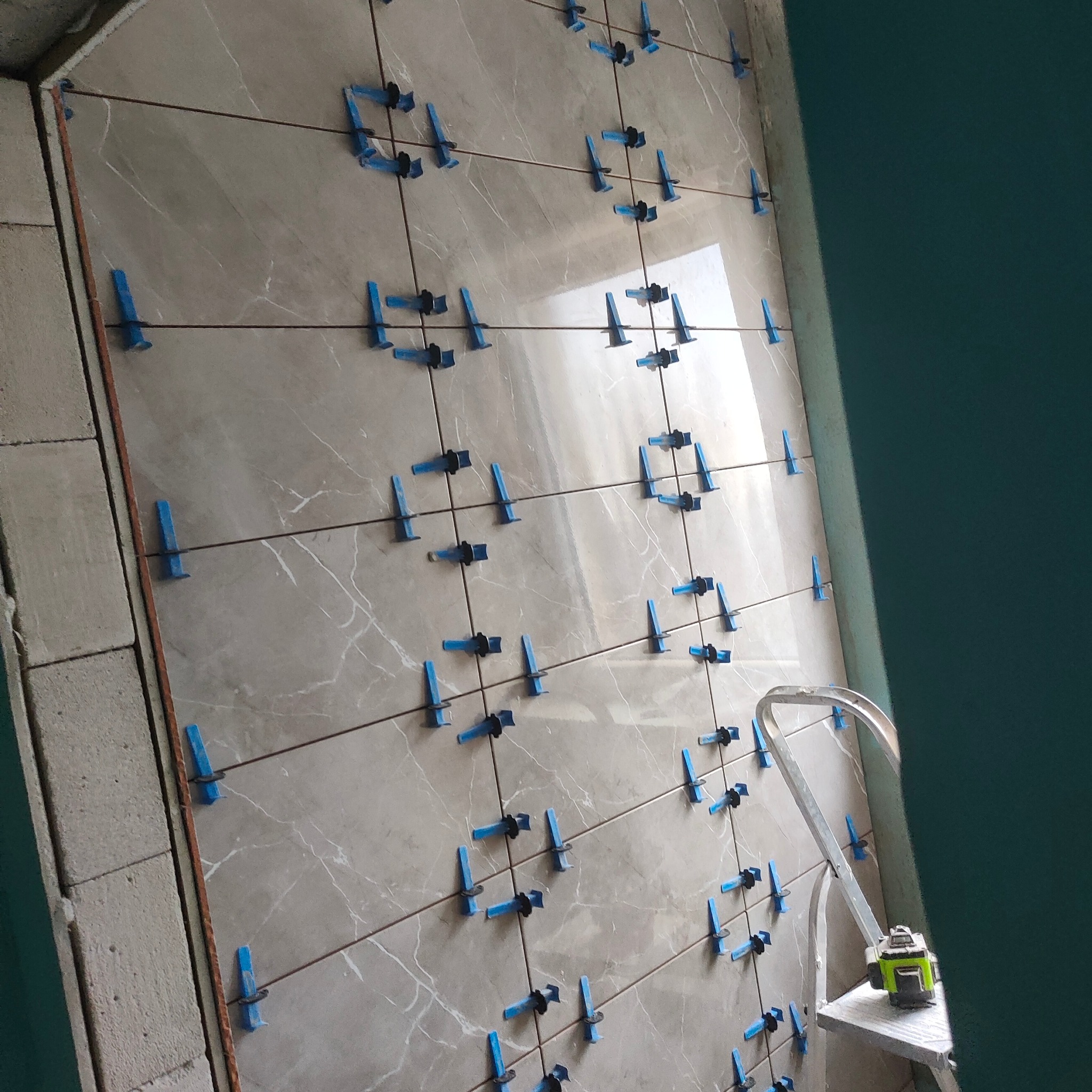 Układanie płytek na ścianie z użyciem poziomicy laserowej i systemu poziomowania. Widoczne krzyżyki dystansowe i kliny. Etap prac wykończeniowych w łazience.