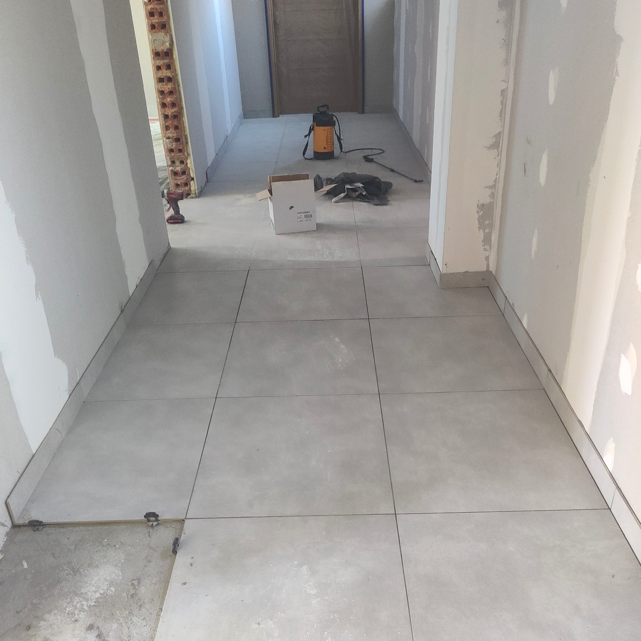 Układanie jasnoszarej, dużej glazury w korytarzu. Widoczne narzędzia i materiały budowlane. Częściowo zakończone prace wykończeniowe ścian.