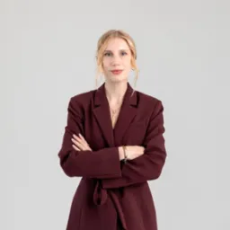 Portret kobiety w bordowej marynarce z paskiem, na jasnym tle, z założonymi rękami. Delikatny uśmiech, blond włosy upięte. Widoczna biżuteria: naszyjnik i bransoletka.