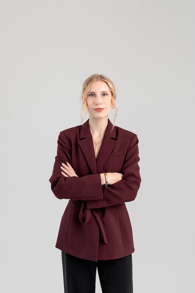 Portret kobiety w bordowej marynarce z paskiem, na jasnym tle, z założonymi rękami. Delikatny uśmiech, blond włosy upięte. Widoczna biżuteria: naszyjnik i bransoletka.