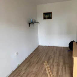 Pokój z panelami podłogowymi w trakcie montażu, widoczne listwy przypodłogowe i fragment mebla. Na ścianie półka i obraz.