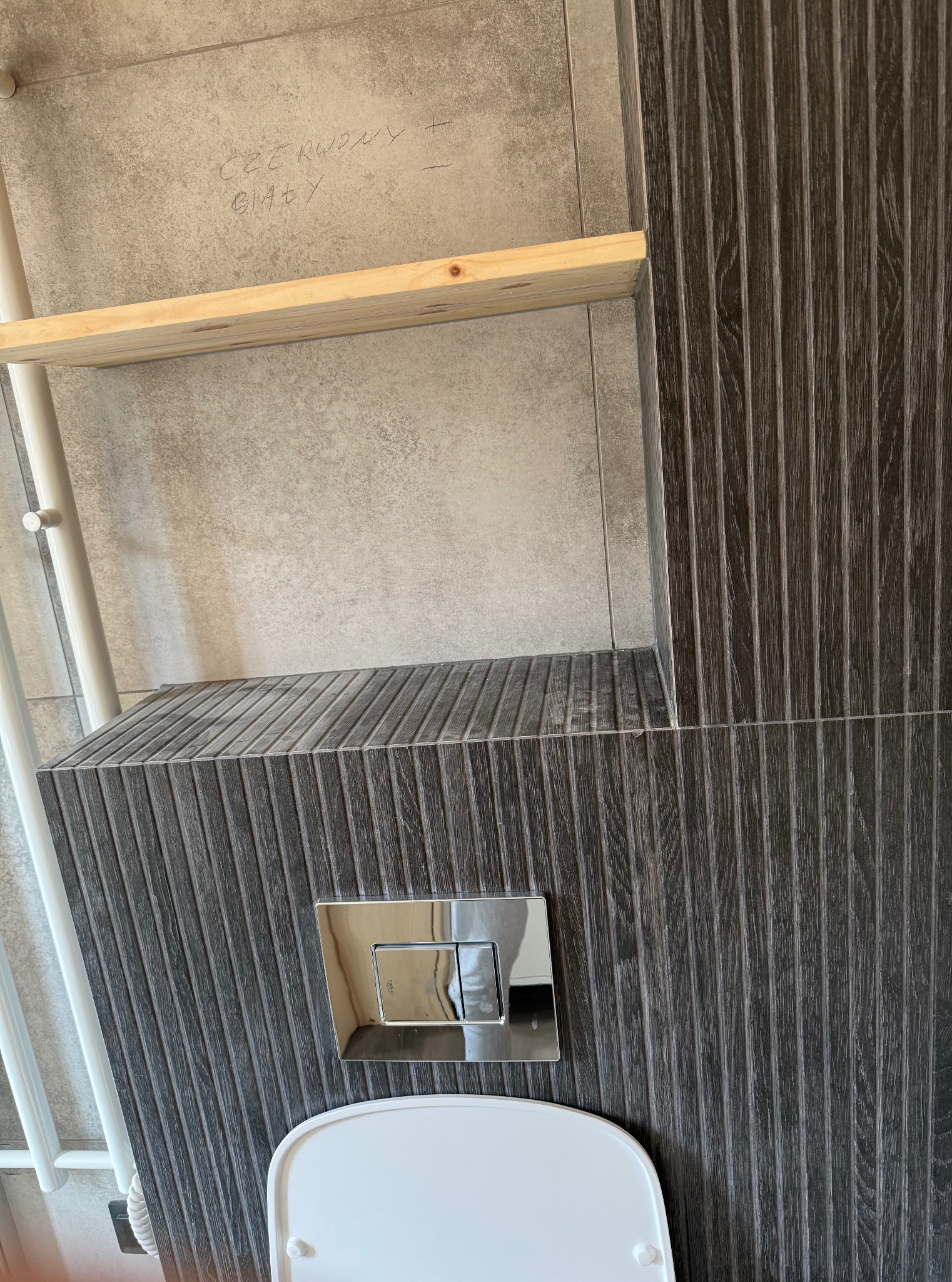 Wykończona łazienka: ciemne, ryflowane płytki imitujące drewno, podwieszana toaleta z chromowanym przyciskiem, drewniana półka i grzejnik drabinkowy.