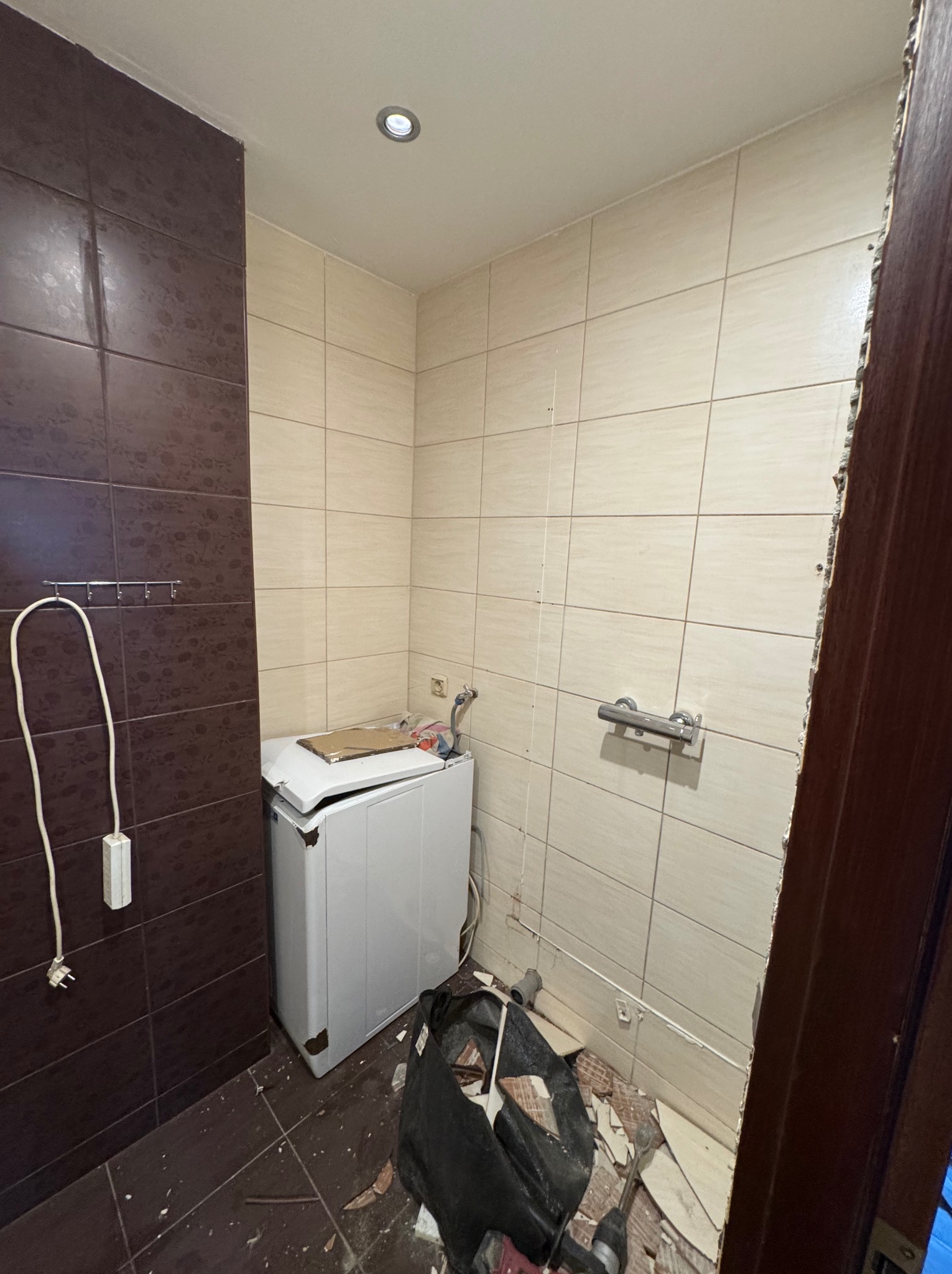 Demontaż płytek w łazience z widoczną pralką i narzędziami. Fragment zdemontowanych płytek podłogowych i ściennych. Etap prac remontowych.