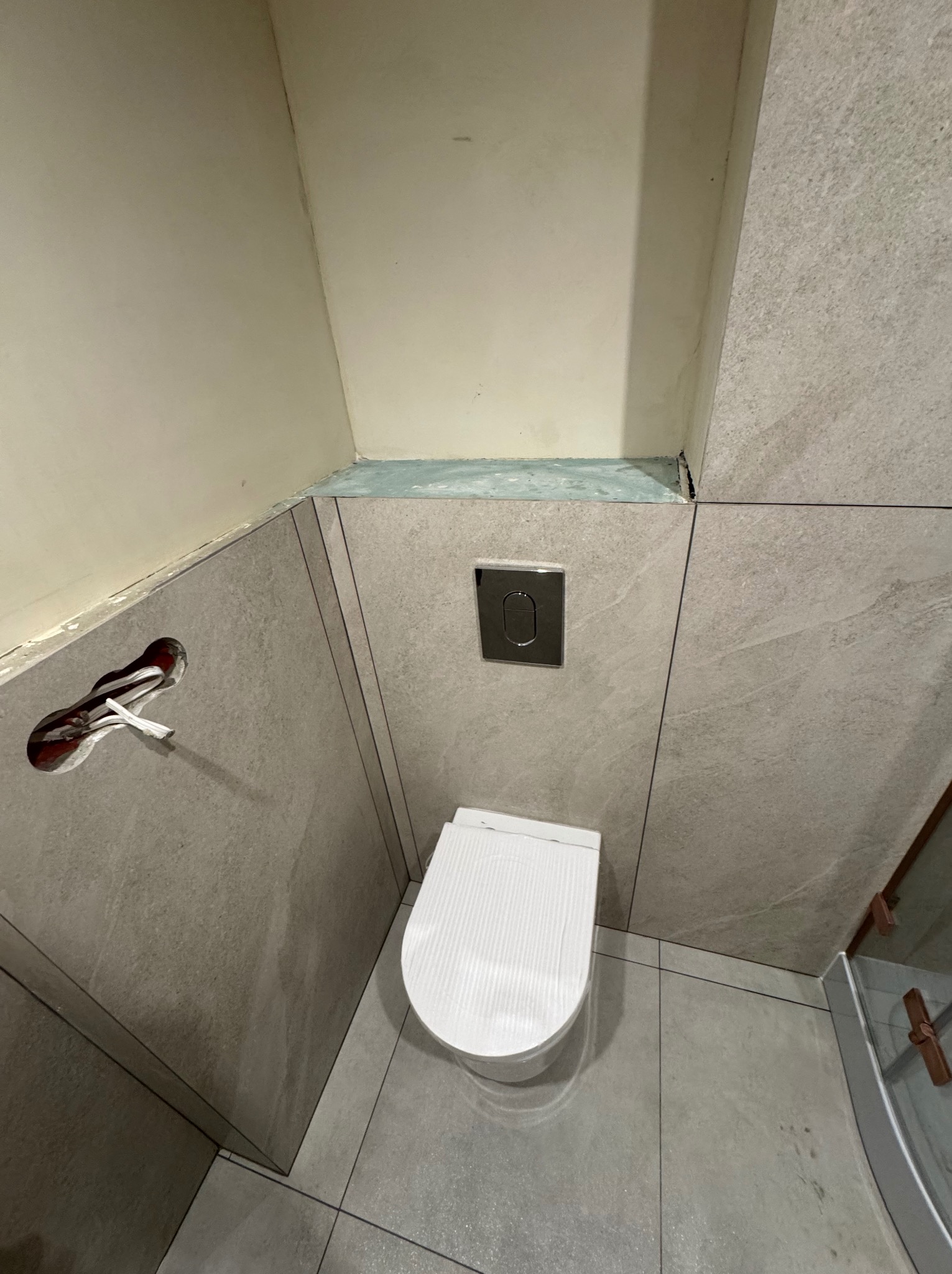 Wykończona toaleta z szarą, kamienną okładziną ścian i podłogi. Widoczny podtynkowy stelaż WC i przycisk spłukujący. W tle fragment kabiny prysznicowej.