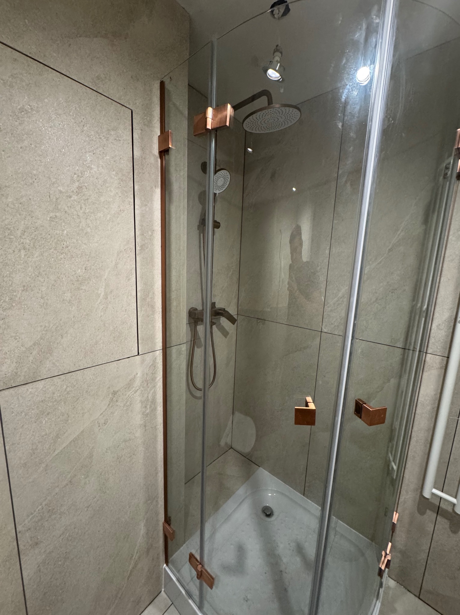 Kabina prysznicowa z miedzianymi detalami w łazience wykończonej płytkami w odcieniach beżu i szarości. Widoczne bateria, deszczownica i odpływ.