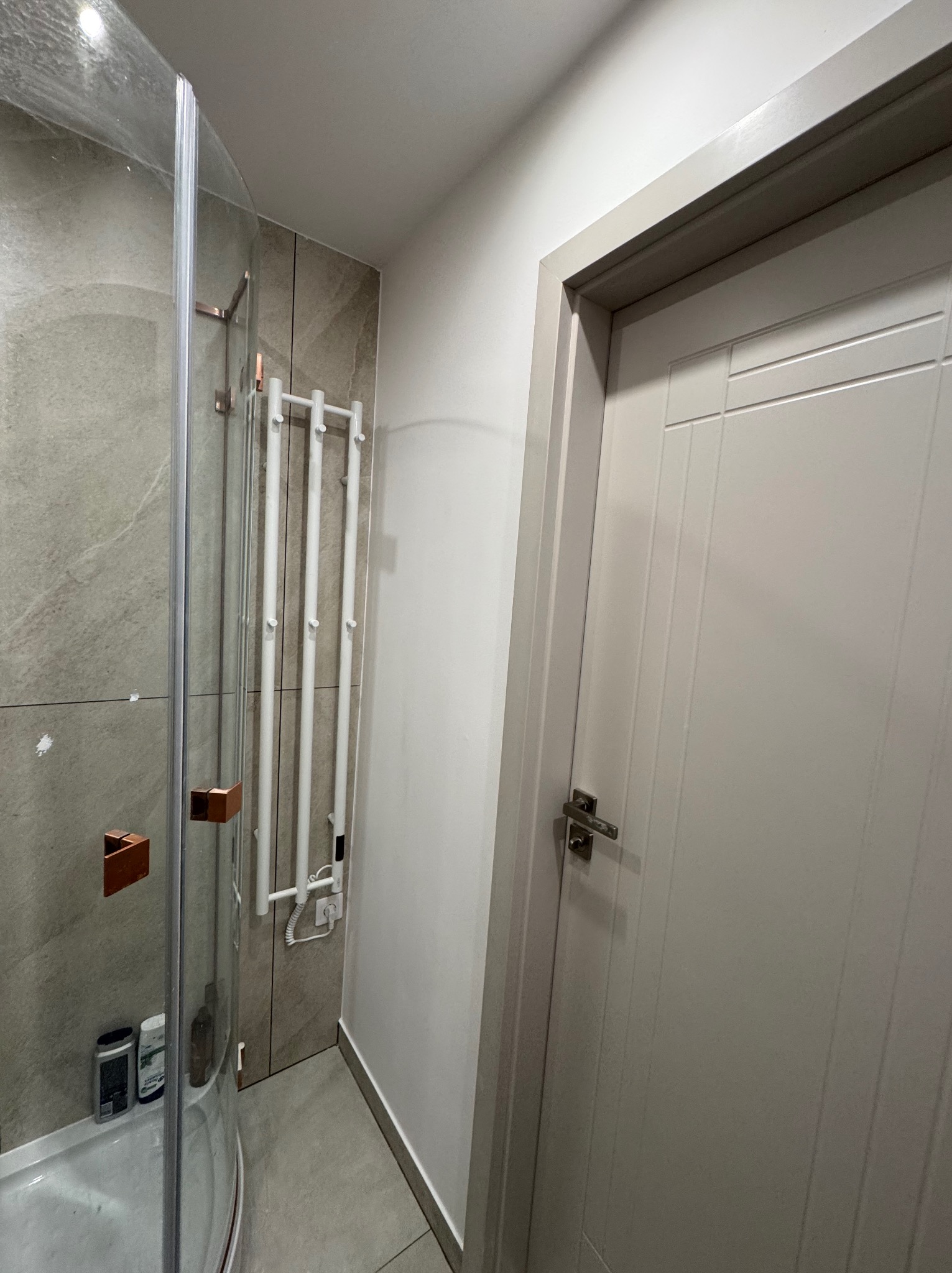 Wykończona łazienka z kabiną prysznicową z zaokrąglonym szkłem i grzejnikiem drabinkowym na tle płytek imitujących kamień. Widoczna szara, panelowa  drzwi.