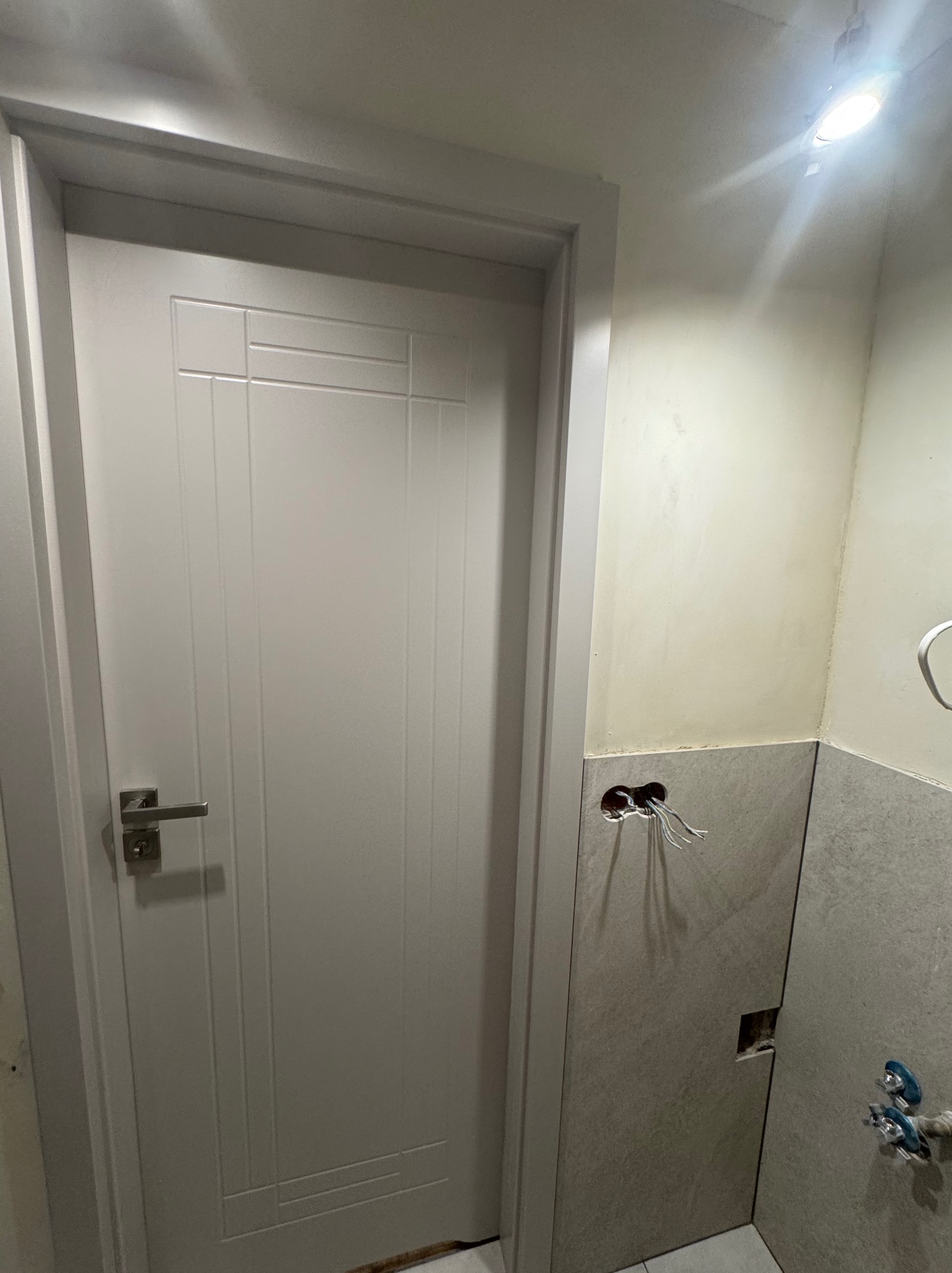 Białe drzwi wewnętrzne z klamką w łazience w trakcie remontu. Widoczne płytki i wystające kable elektryczne.