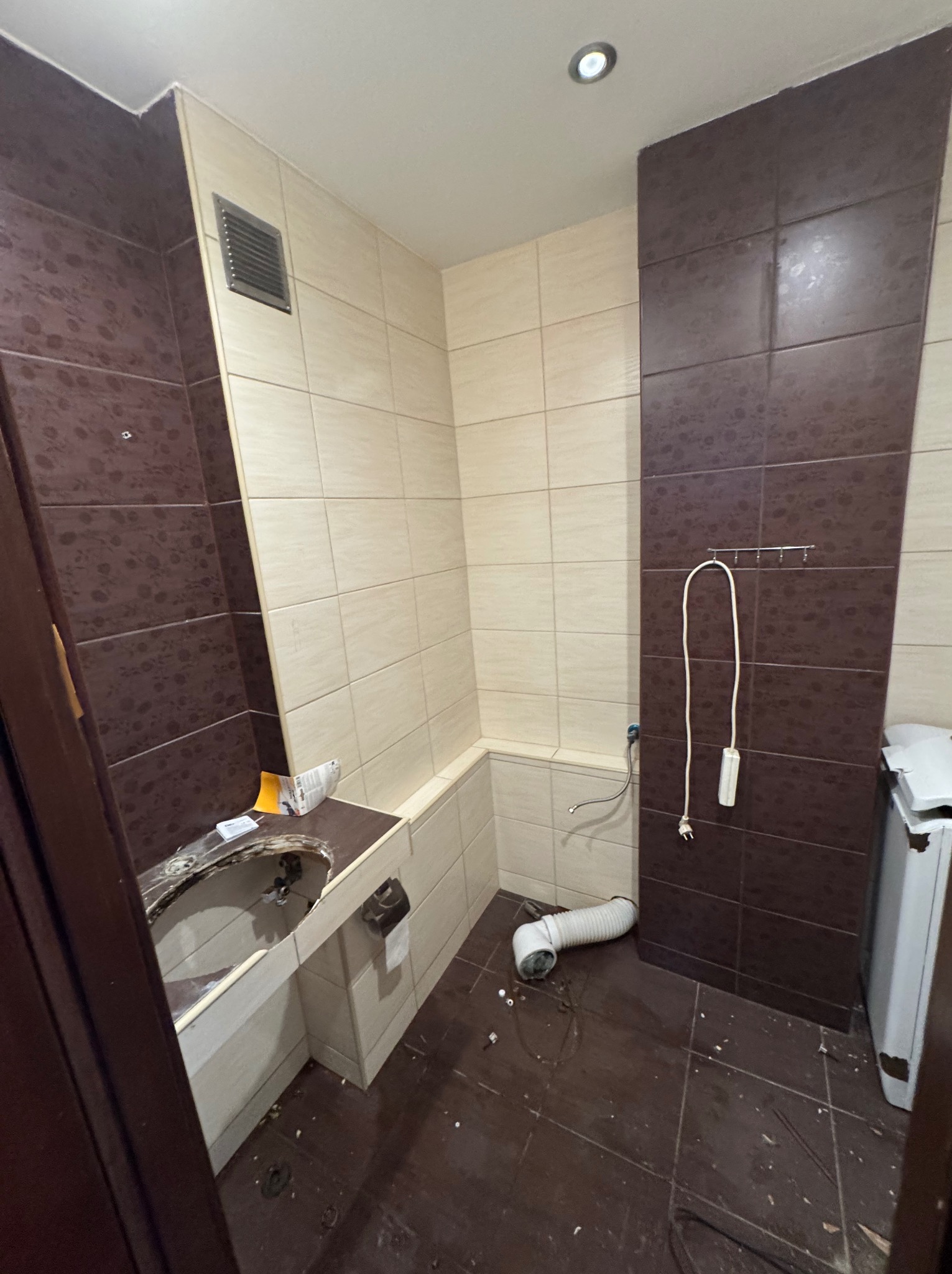 Łazienka w trakcie prac: brązowe i beżowe płytki, fragment umywalki bez armatury, widoczne rury i akcesoria. Częściowy demontaż wyposażenia.