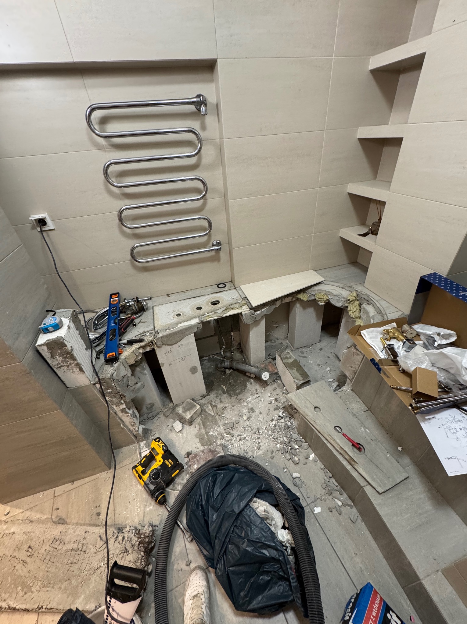 Wnętrze łazienki w trakcie prac remontowych: usunięte płytki podłogowe, widoczne narzędzia, grzejnik drabinkowy na ścianie, elementy instalacji. Fragmentaryczny widok.