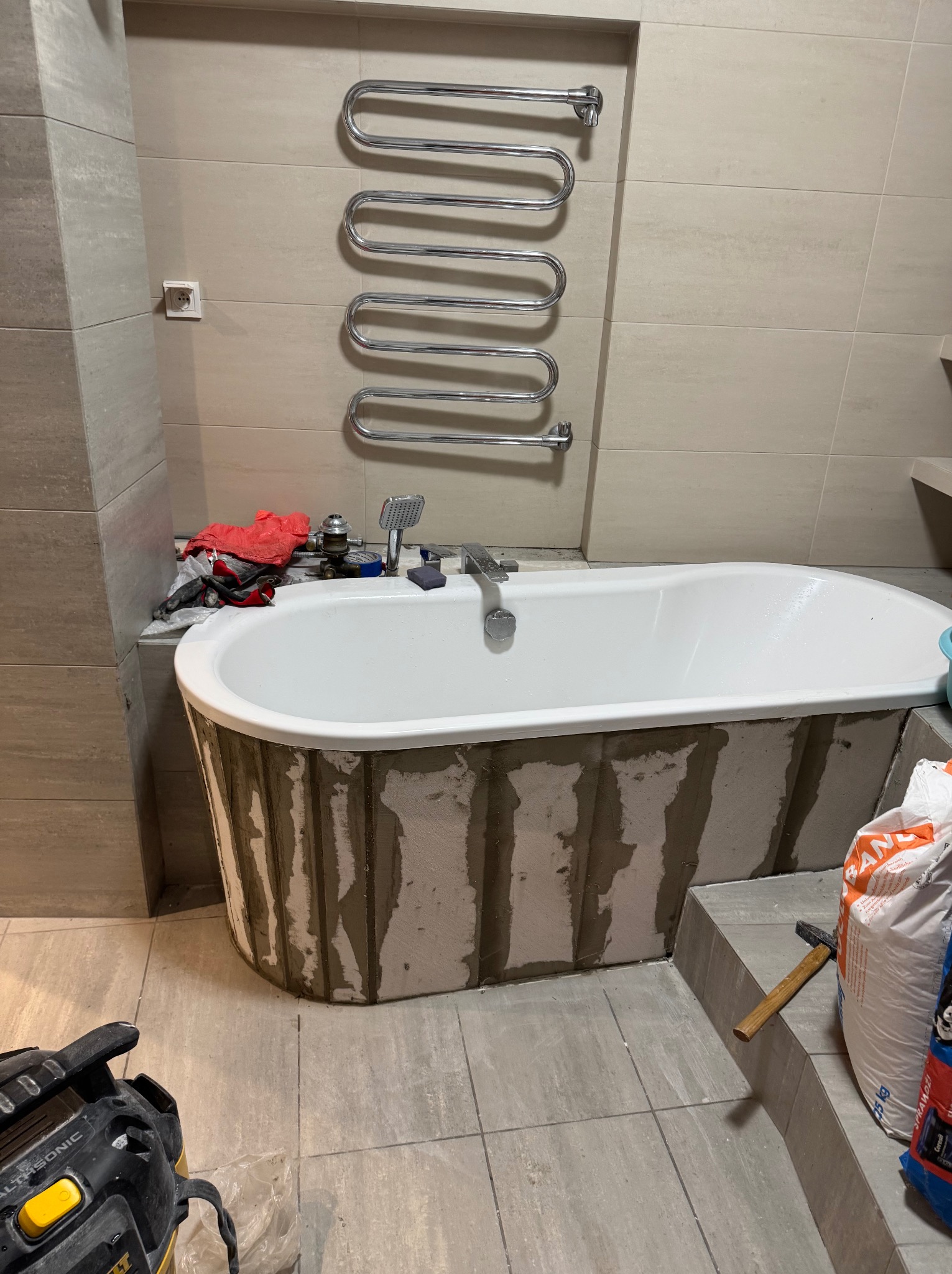 Biała wanna z obudową w trakcie wykańczania, widoczny grzejnik drabinkowy i narzędzia. Szare płytki na ścianach i podłodze. Prace wykończeniowe w łazience.