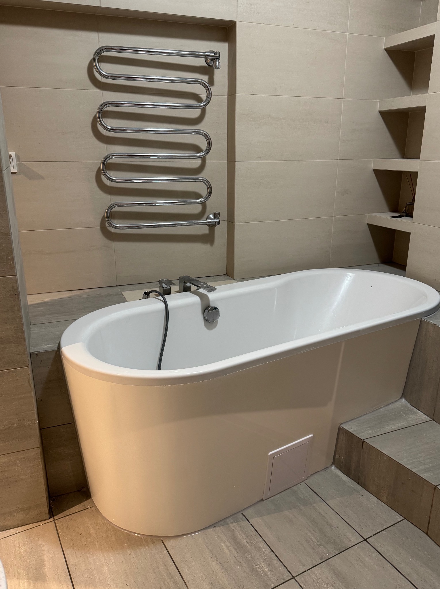 Biała wanna wolnostojąca z chromowaną armaturą na tle beżowych płytek. Na ścianie grzejnik drabinkowy i półki wnękowe.