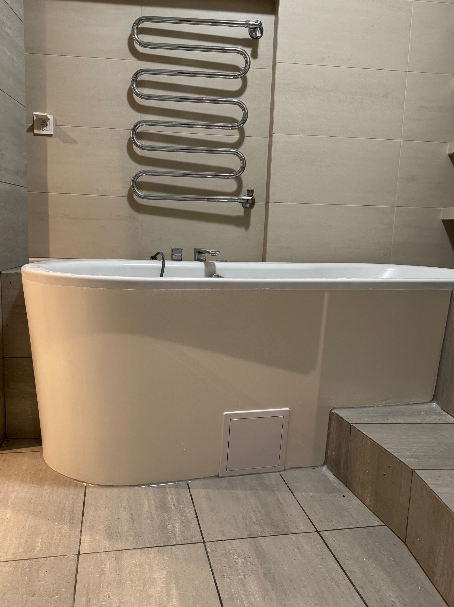 Nowoczesna łazienka: beżowe płytki, wanna z obudową i grzejnik drabinkowy. Widoczny fragment podłogi i gniazdko elektryczne.