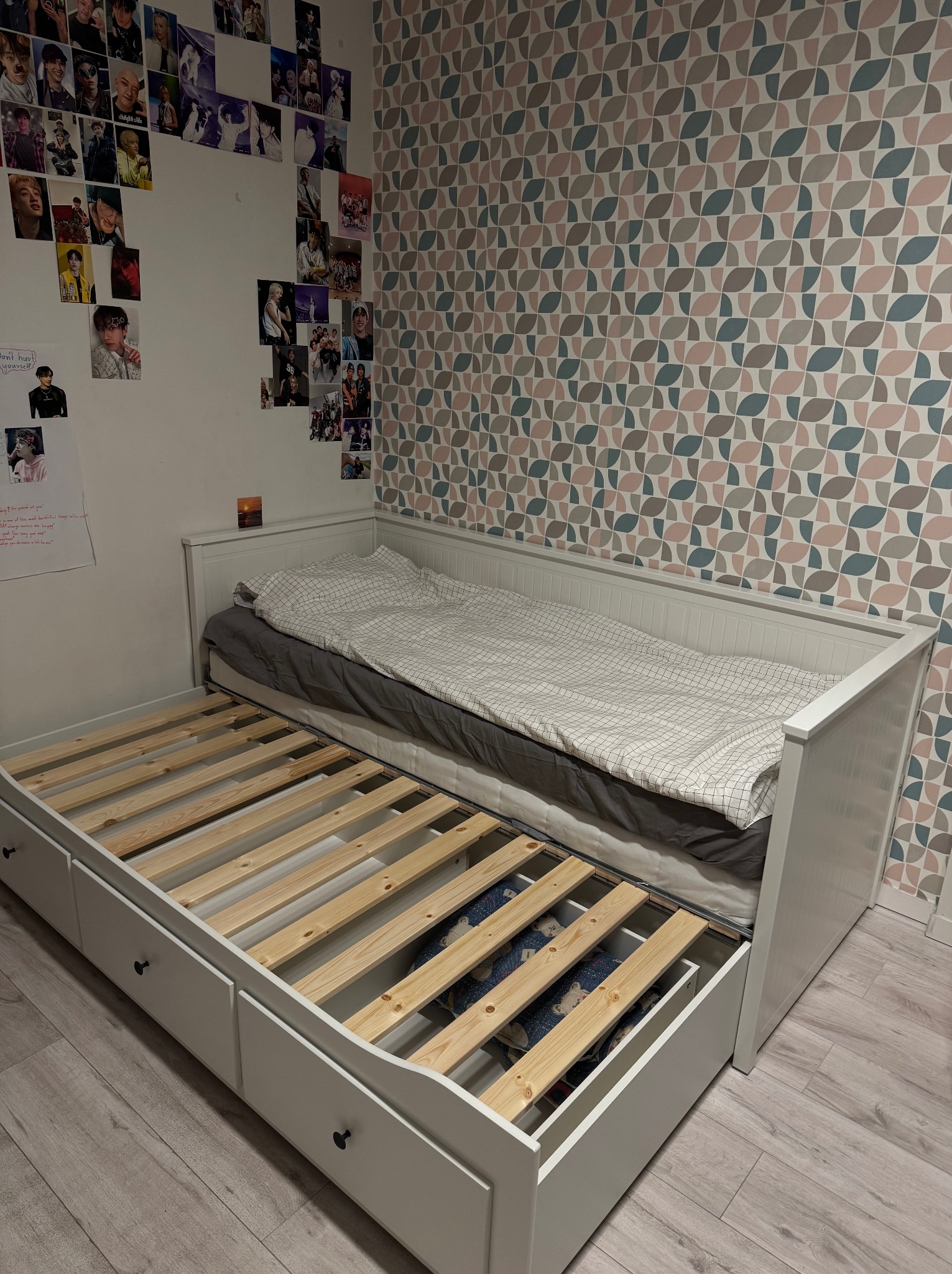 Rozłożone białe łóżko z wysuwaną częścią, widoczne drewniane stelaże i szuflady. W tle ściana z wzorzystą tapetą i plakatami.