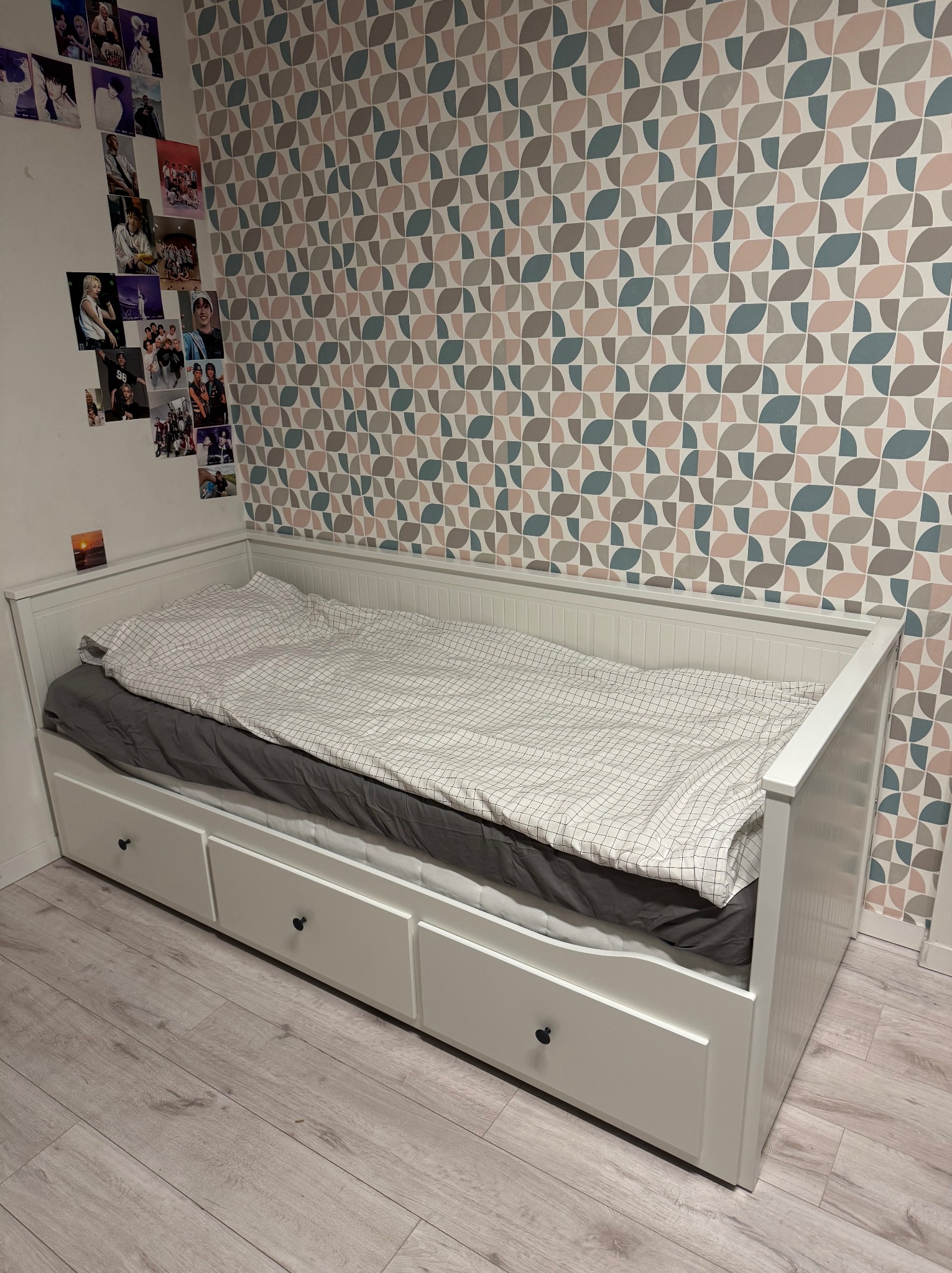 Białe łóżko z szufladami w pokoju z wzorzystą tapetą i zdjęciami na ścianie. Widok na podłogę z paneli i fragment wnęki okiennej.