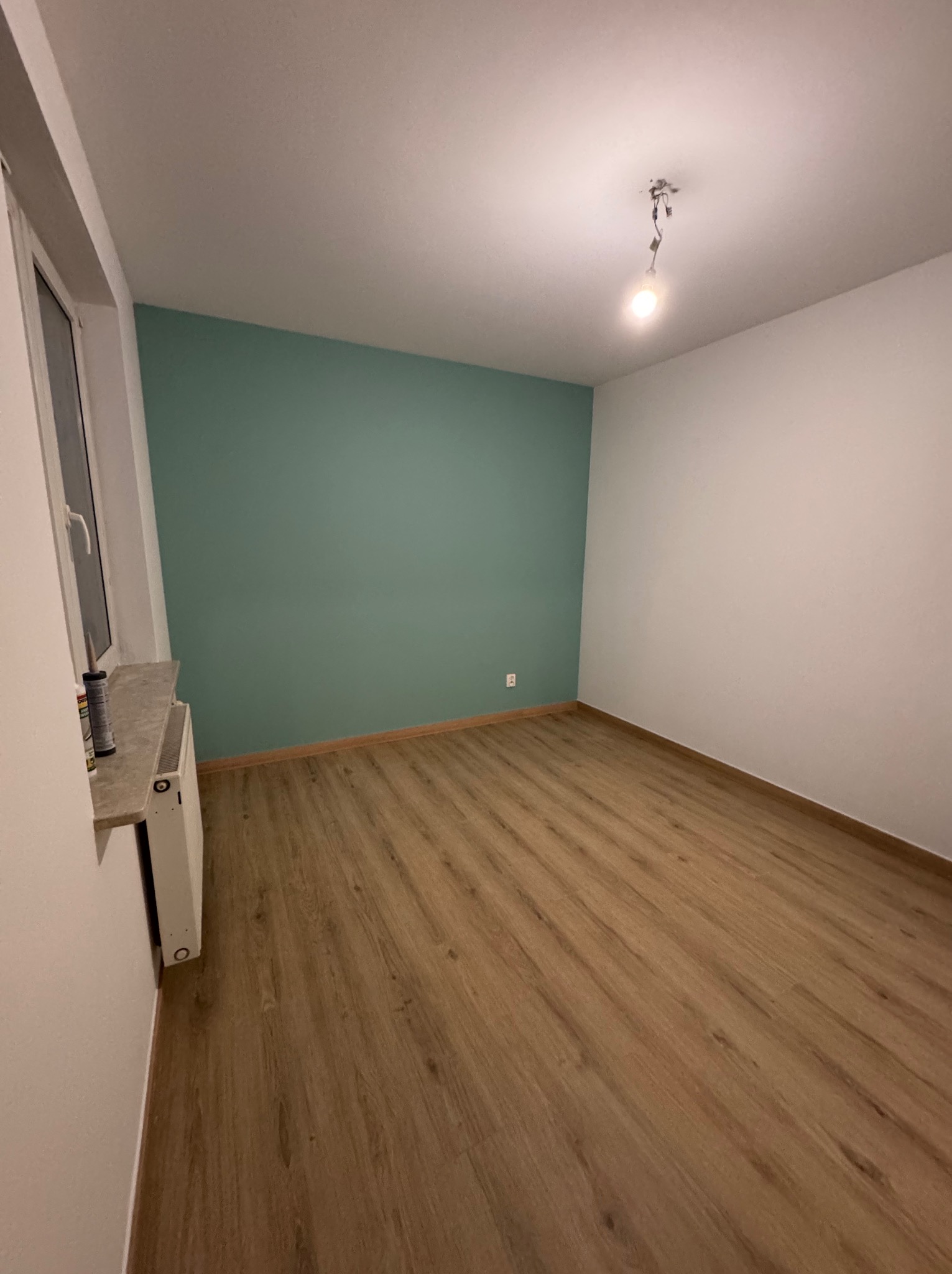 Pokój po remoncie: świeżo położone panele podłogowe w odcieniach drewna, pomalowana na zielono ściana, białe ściany, okno z parapetem. Na suficie wisi żarówka.