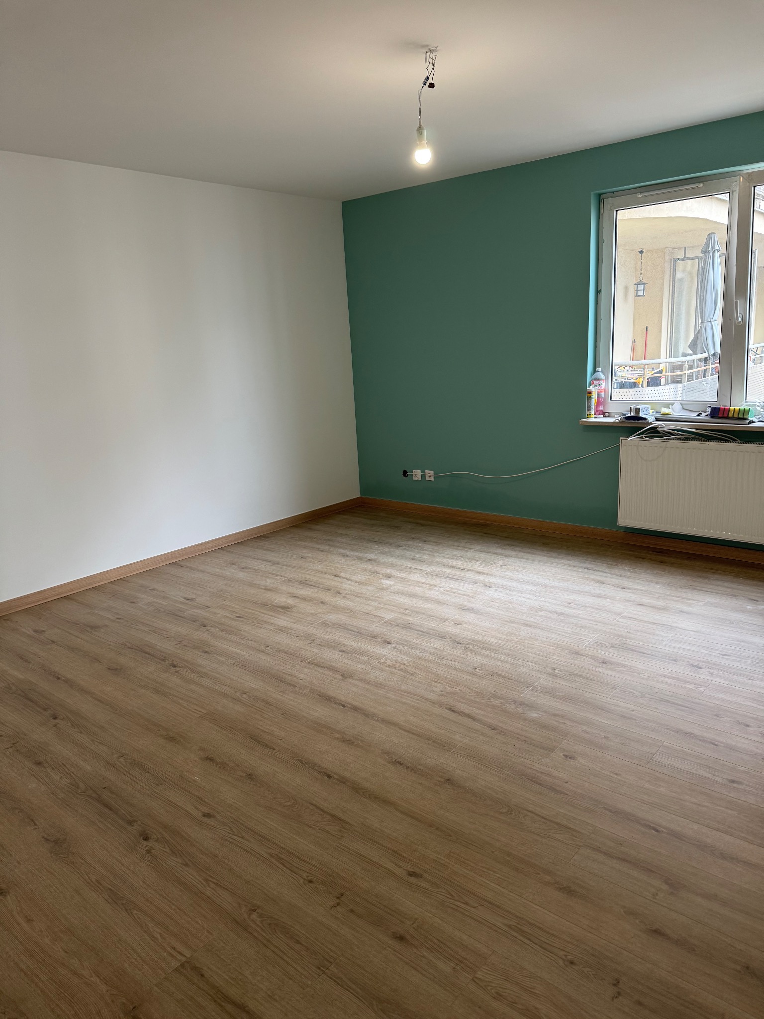 Pomalowany pokój z panelami podłogowymi w odcieniach brązu. Jedna ściana biała, druga turkusowa. Widoczne gniazdka elektryczne i grzejnik pod oknem.