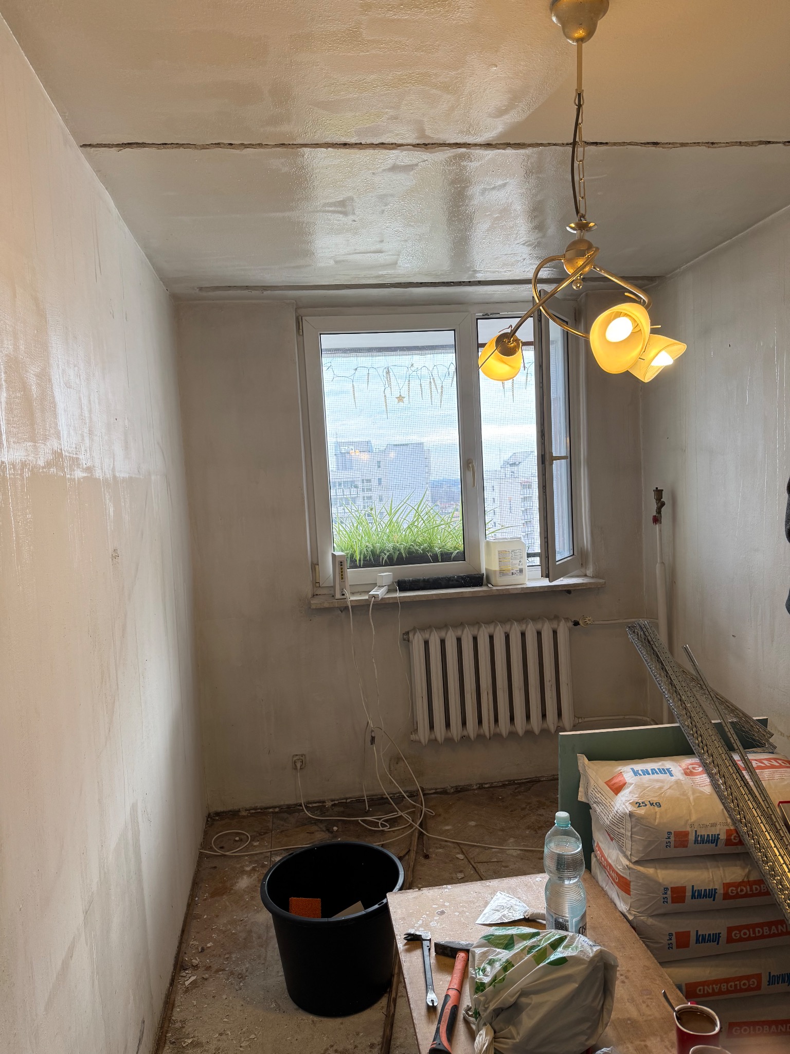 Pokój w trakcie szpachlowania ścian i sufitu, widoczne worki z gładzią, narzędzia, okno z widokiem na miasto. Wnętrze w trakcie prac wykończeniowych.