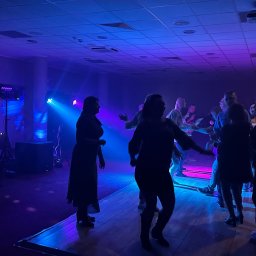 Grupa wodzirejsko - DJ GK - Tańczący ludzie na parkiecie oświetlonym niebieskimi i fioletowymi światłami. Widoczne nagłośnienie i oświetlenie sceniczne. Impreza w lokalu.