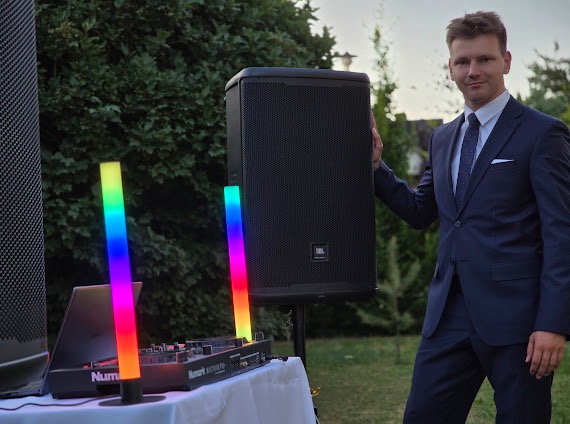 Mężczyzna w garniturze prezentuje sprzęt DJ-ski: mikser Numark, głośniki JBL i kolorowe oświetlenie LED na tle zieleni. Scena gotowa do imprezy.