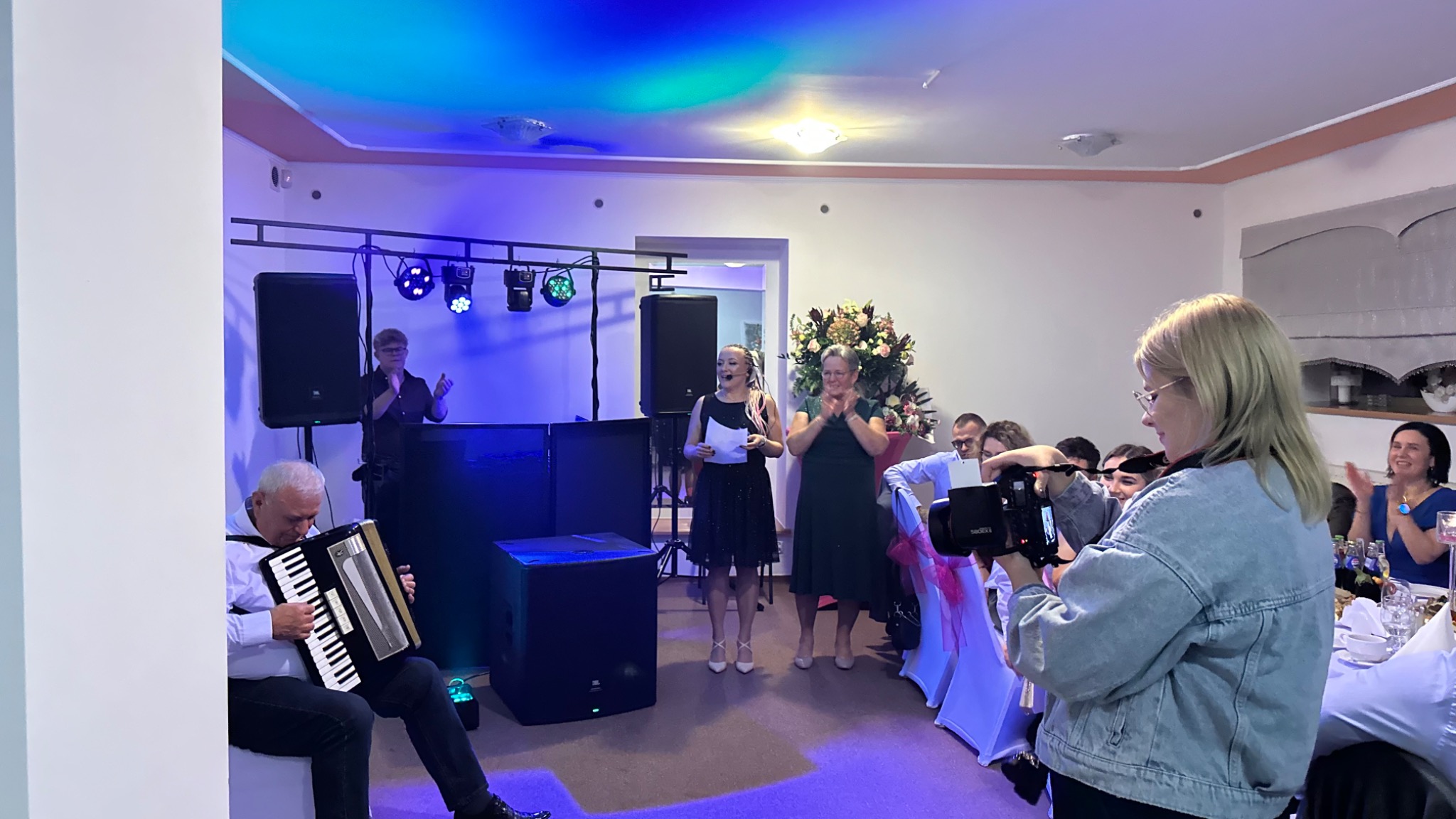 Zespół muzyczny gra na akordeonie podczas imprezy, oświetlenie sceniczne, DJ za konsoletą, konferansjerka prowadzi wydarzenie, fotograf uwiecznia momenty.