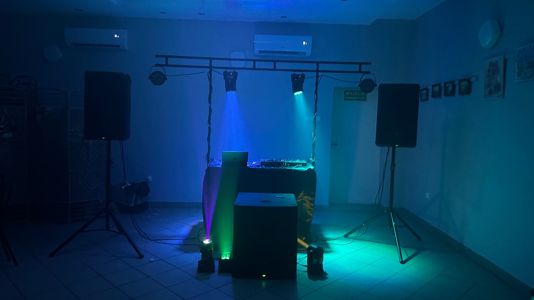 Sprzęt nagłośnieniowy i oświetleniowy z konsoletą DJ-ską w pomieszczeniu, oświetlony niebiesko-zielonym światłem. Widoczne głośniki na statywach i reflektory.