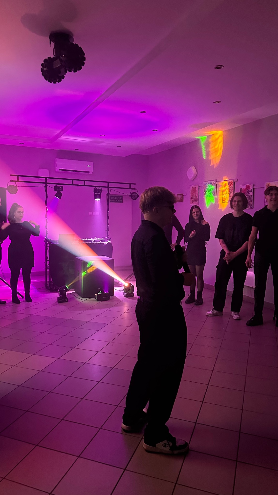 Oświetlenie sceniczne w fiolecie, DJ-ka i grupa osób na imprezie. Reflektory tworzą kolorowe efekty świetlne. Widoczny parkiet i elementy wyposażenia sali.