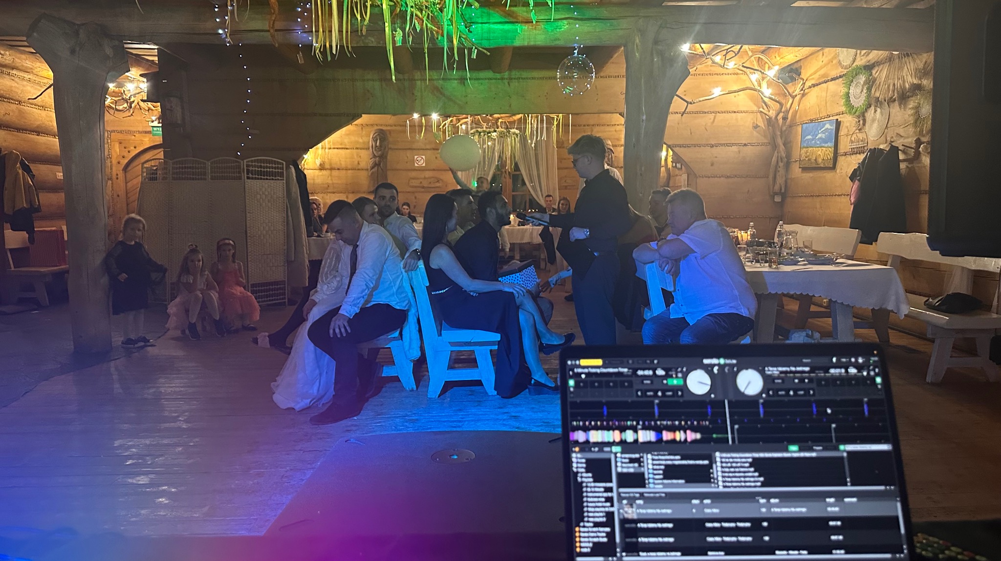DJ obsługuje wesele w drewnianej sali. W tle goście siedzą na krzesłach, a kelner podaje posiłki. Na pierwszym planie widoczny ekran laptopa z oprogramowaniem DJ-skim.