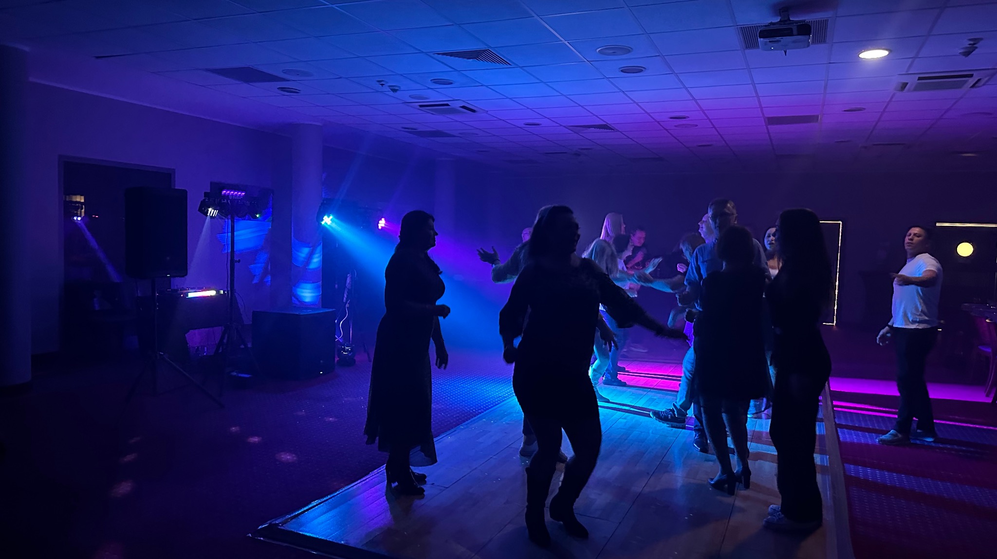 Tańczący ludzie na parkiecie oświetlonym niebieskimi i fioletowymi światłami. Widoczne nagłośnienie i oświetlenie sceniczne. Impreza w lokalu.