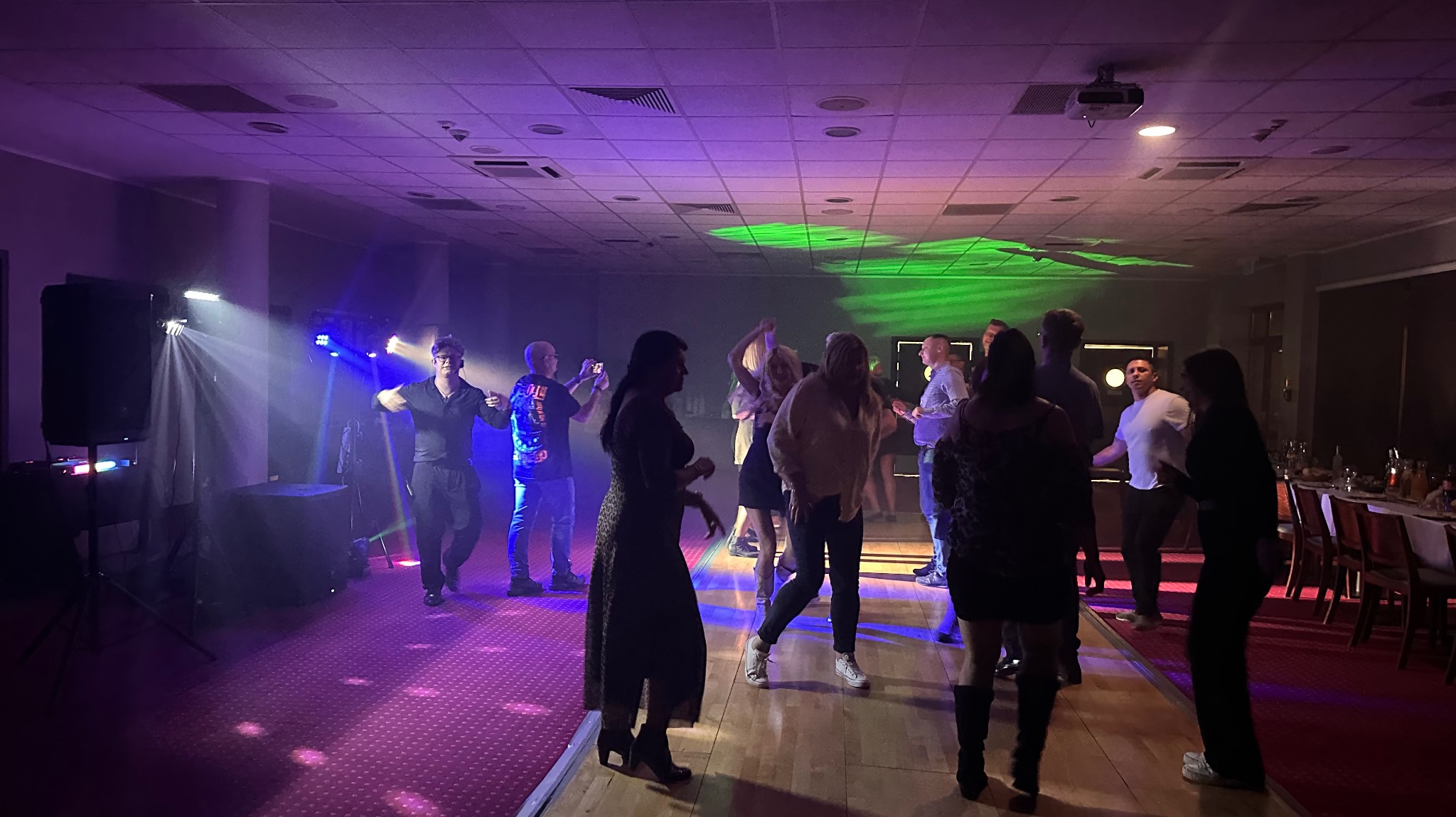 Zabawa taneczna w sali z oświetleniem eventowym: reflektory, parkiet, bawiący się ludzie. Widoczne stoły z zastawą w tle. Dynamiczne ujęcie z imprezy.