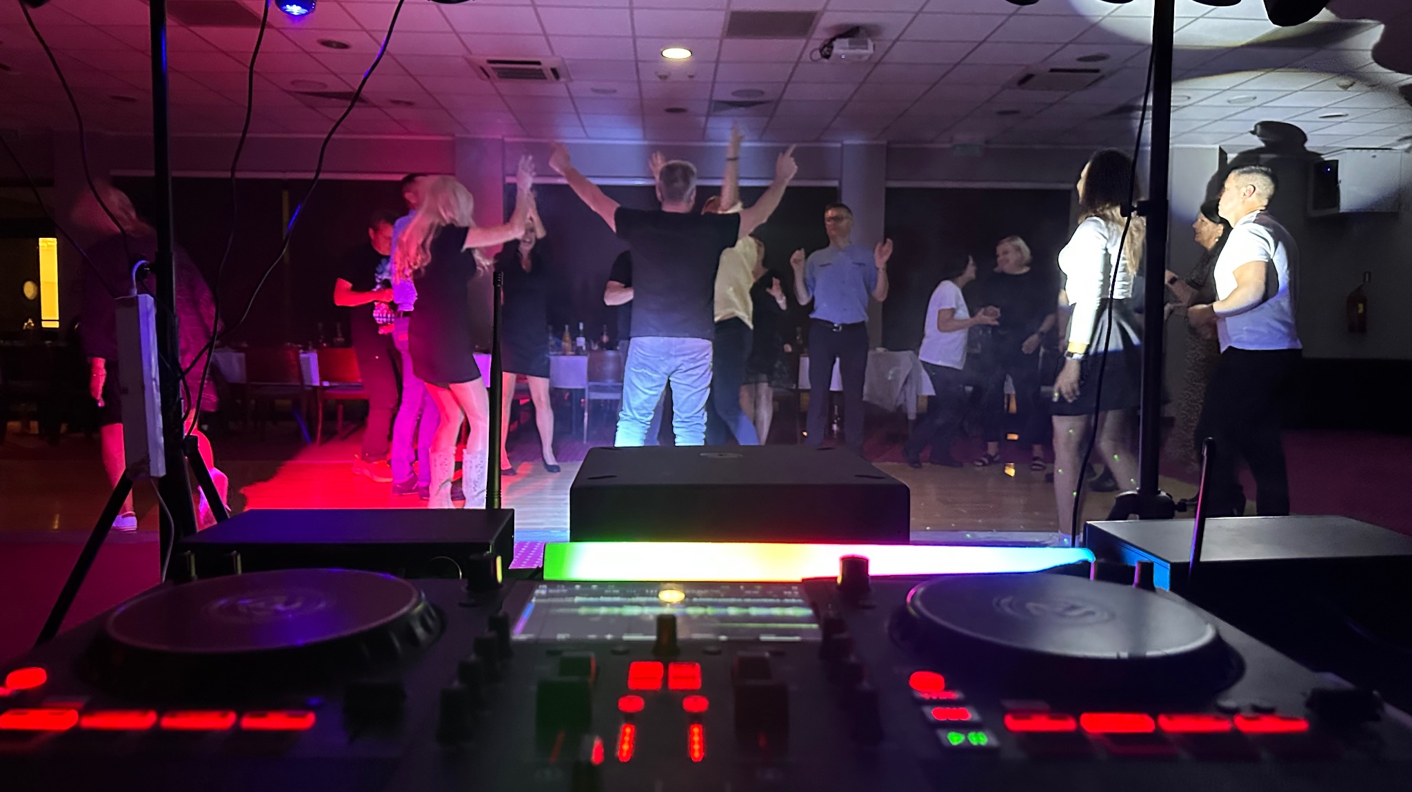 Parkiet taneczny z bawiącymi się ludźmi, oświetlony kolorowymi światłami, z widocznym sprzętem DJ-skim na pierwszym planie. Dynamiczna atmosfera imprezy.