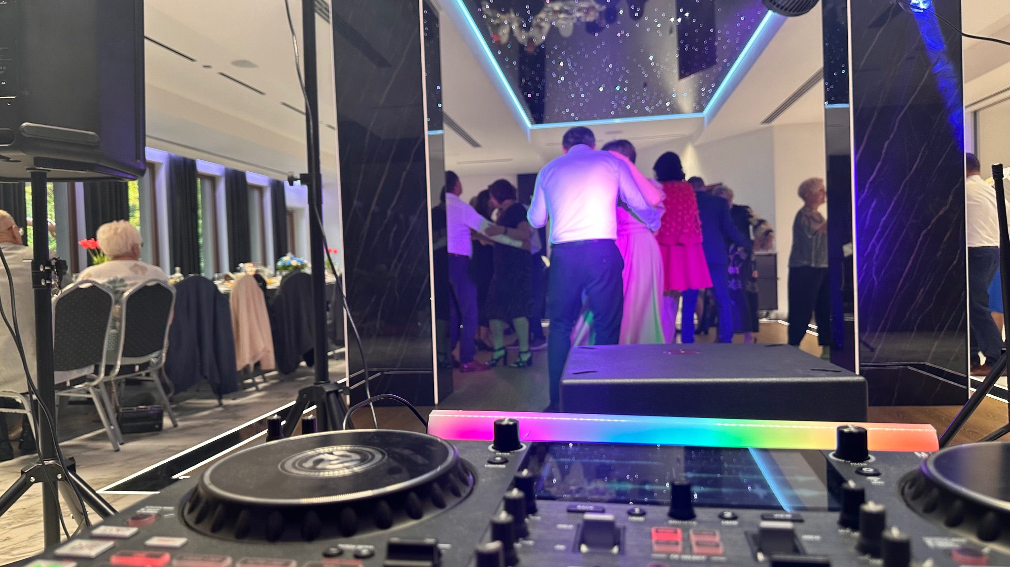DJ na weselu: mikser z tęczowym oświetleniem na pierwszym planie, tańczący goście w tle, elegancka sala z lustrami i dekoracyjnym oświetleniem sufitowym.