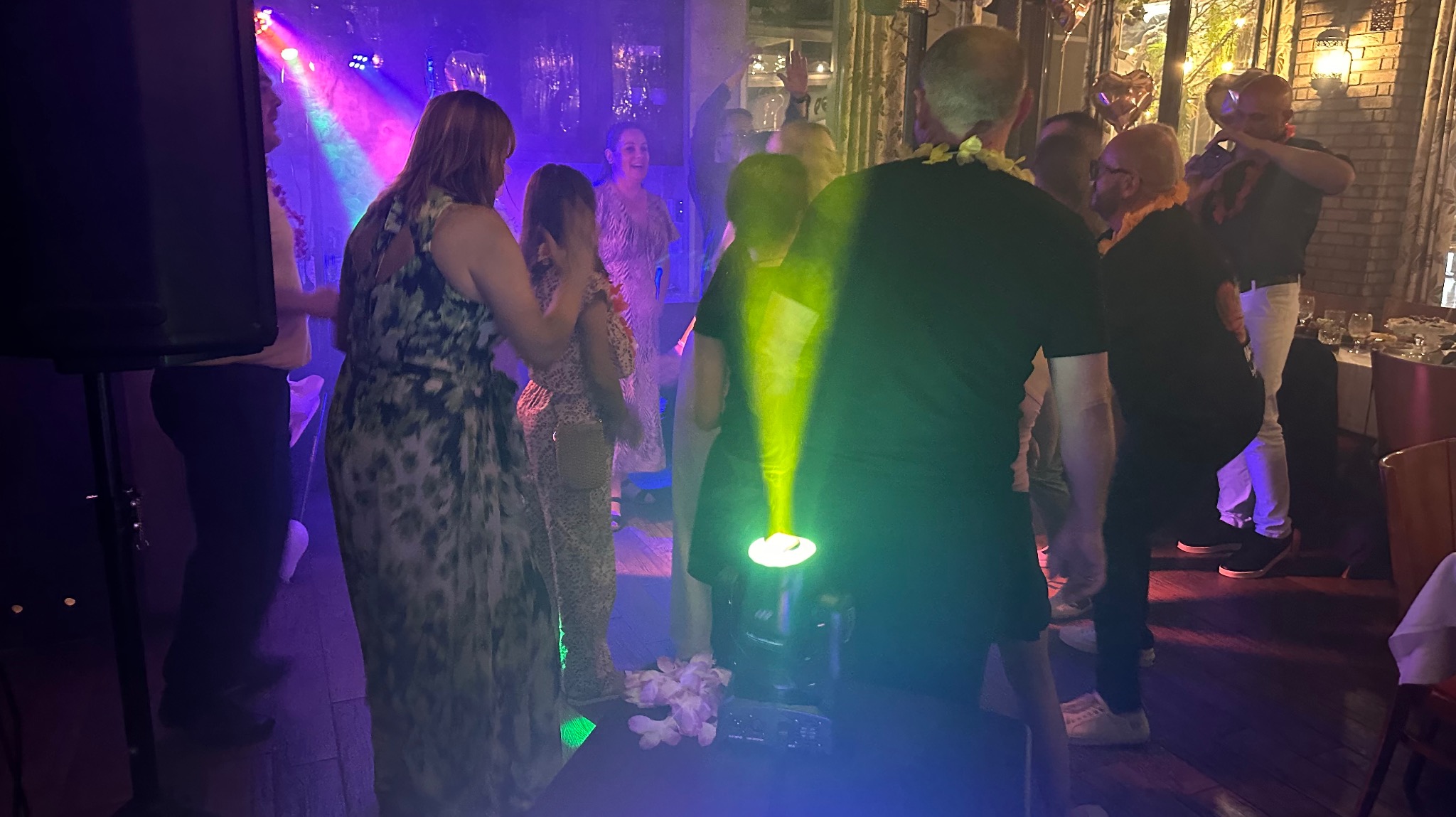 Impreza w stylu hawajskim z oświetleniem scenicznym. Goście w kwiatowych lei tańczą w blasku reflektorów. Widoczne stoły z nakryciami i dekoracje.