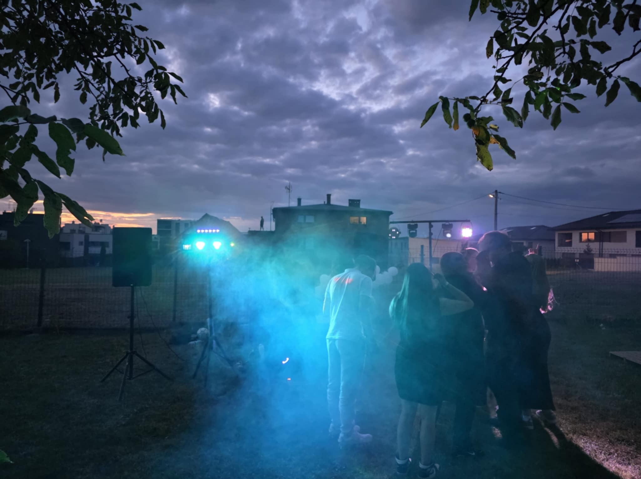 Wieczorny plener z nagłośnieniem i oświetleniem podczas imprezy. W tle budynki i zachmurzone niebo. W kadrze osoby w dymie i sprzęt nagłośnieniowy.