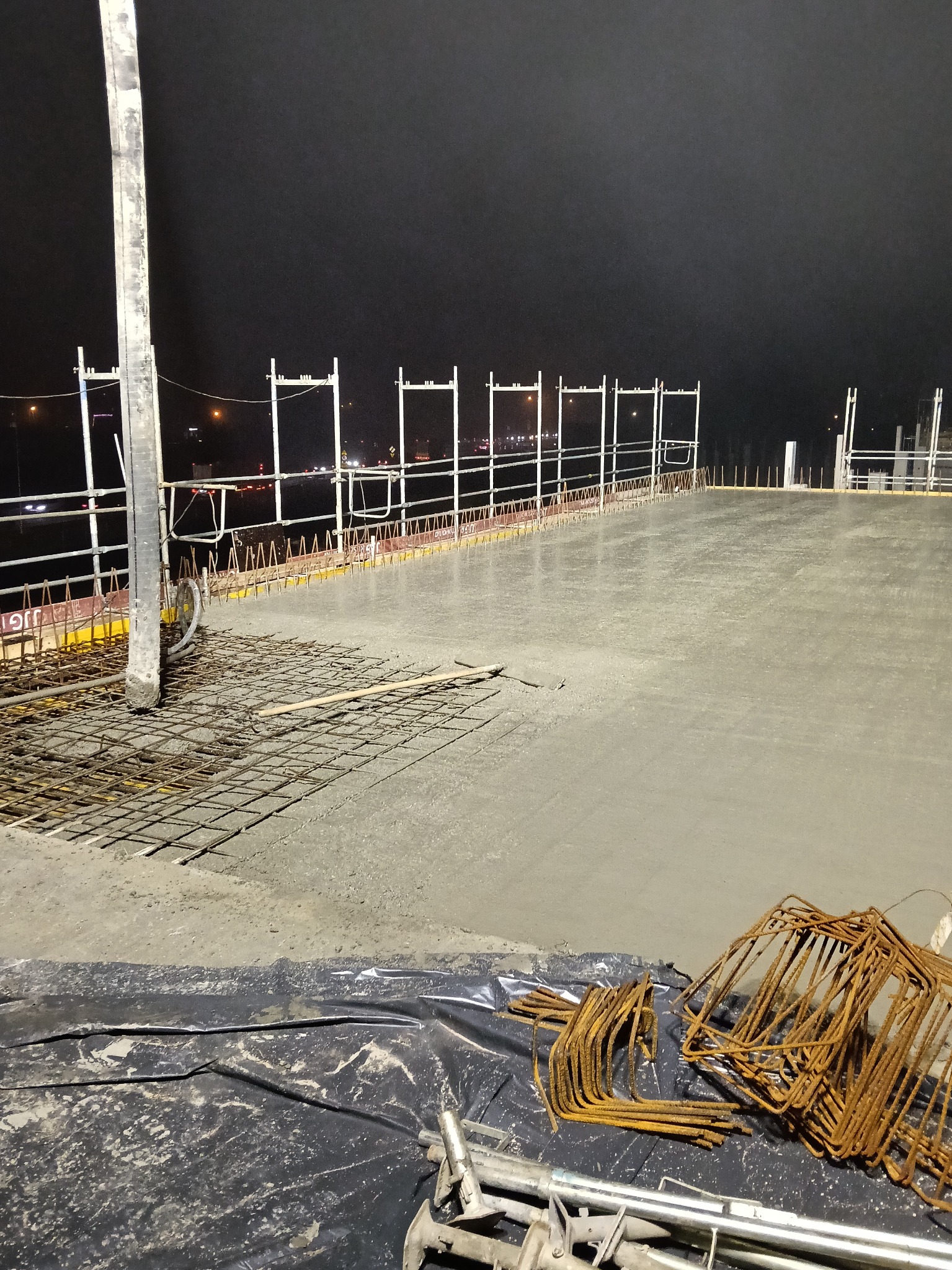 Zalewanie betonem zbrojonej płyty stropowej na budowie nocą. Widoczne szalunki, wystające pręty i pompa do betonu. Częściowo zalana powierzchnia, zbrojenie i narzędzia.