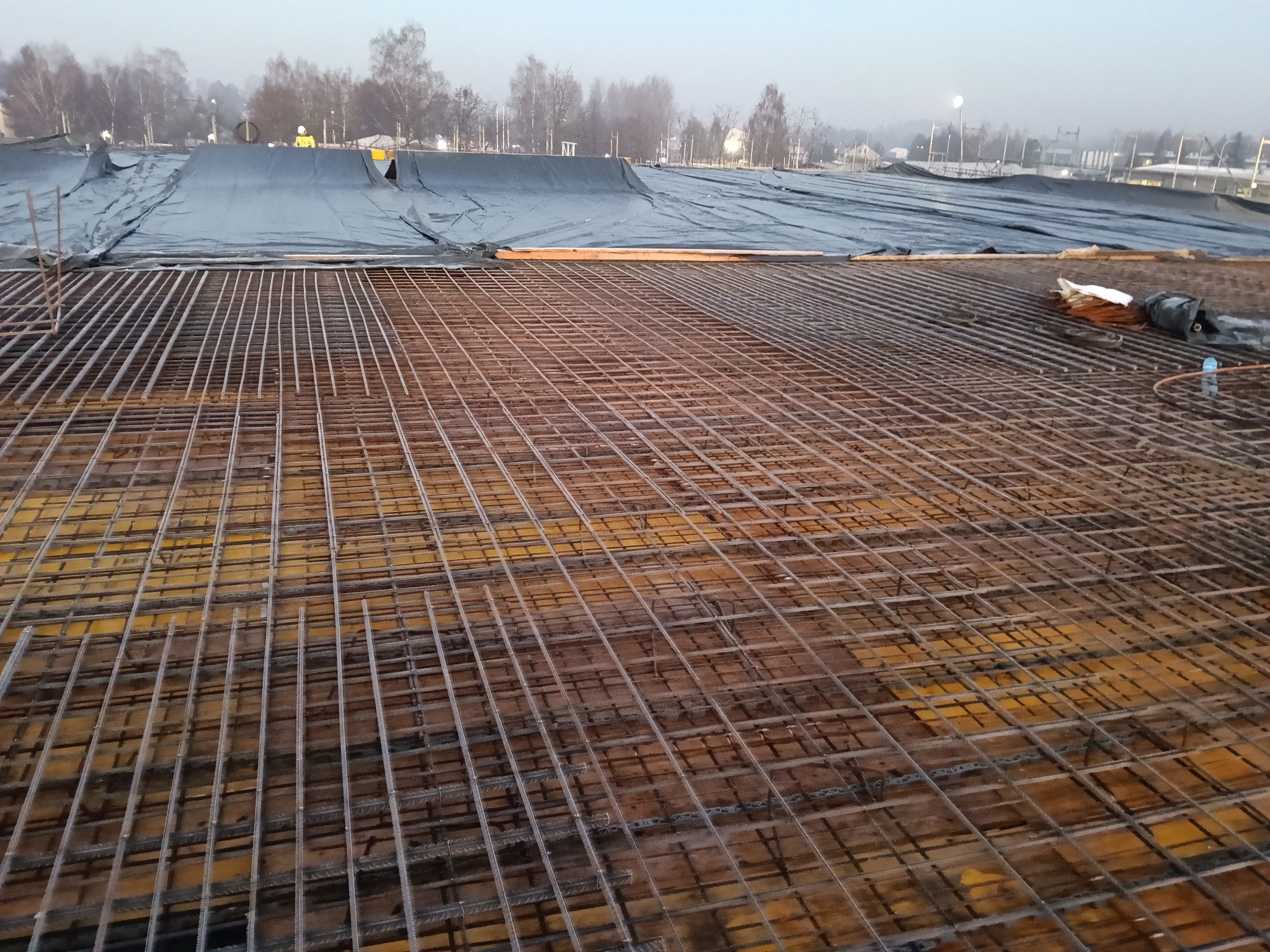Siatka zbrojeniowa na deskowaniu, przygotowana do zalania betonem. W tle widoczna plandeka i zarys zabudowań w oddali, w pochmurny dzień.