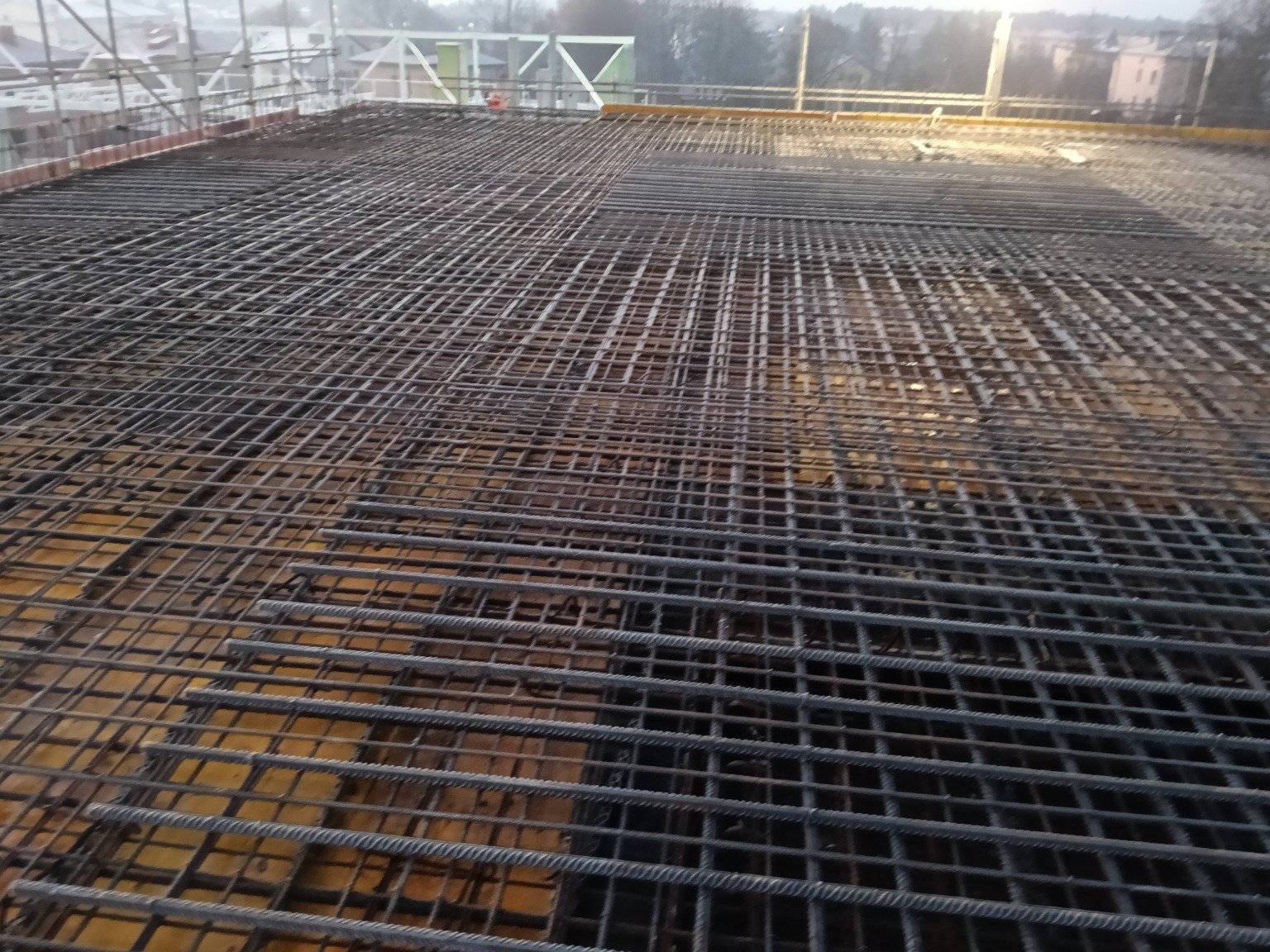 Siatka zbrojeniowa ułożona na szalunku, przygotowana do zalania betonem. Widok z góry, zarys budynków w tle. Zbrojenie na budowie w pochmurny dzień.