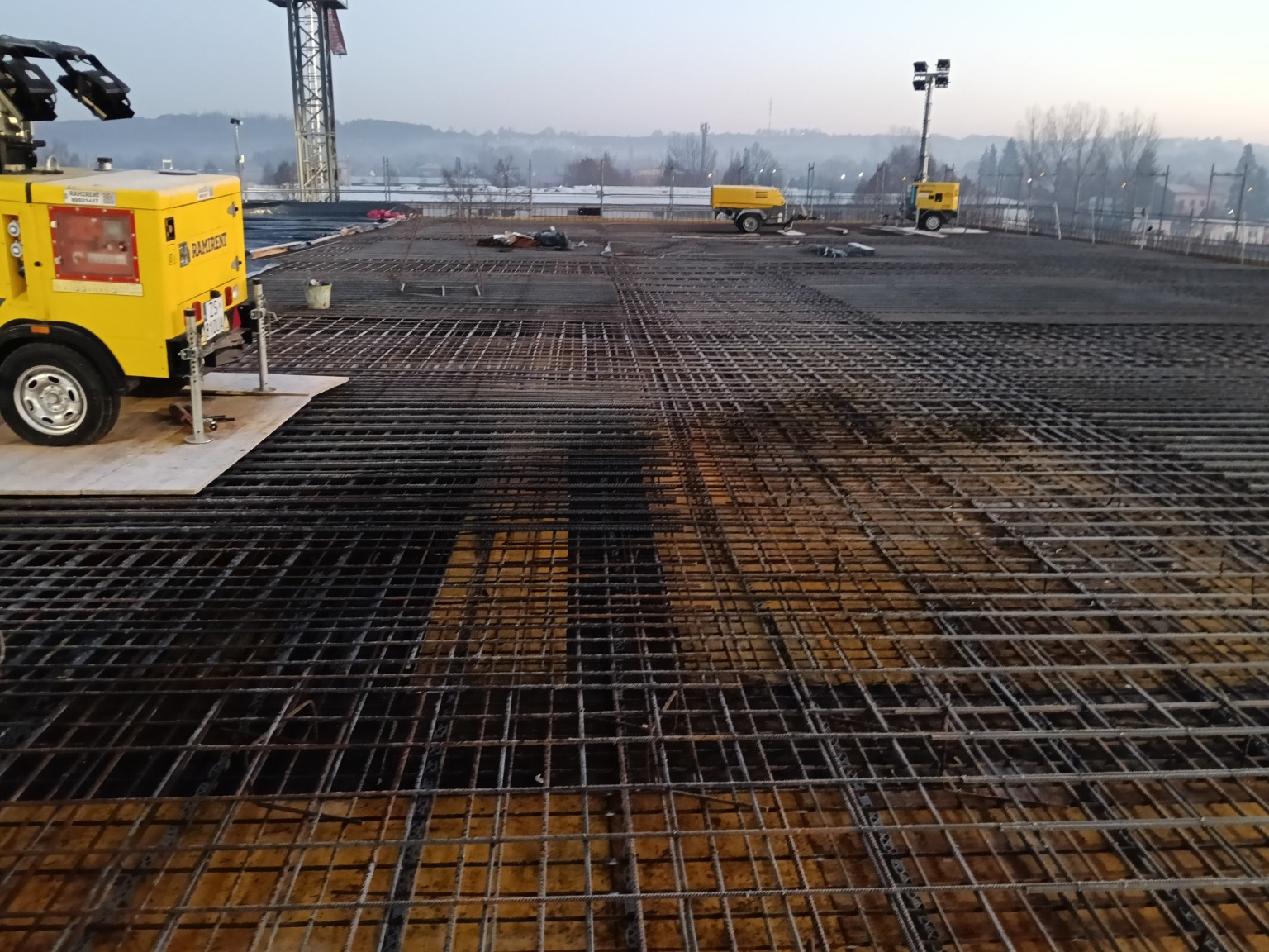 Siatka zbrojeniowa na budowie, z żółtymi agregatami prądotwórczymi w tle, wczesnym rankiem. Widoczne elementy konstrukcji i krajobraz miejski w oddali.