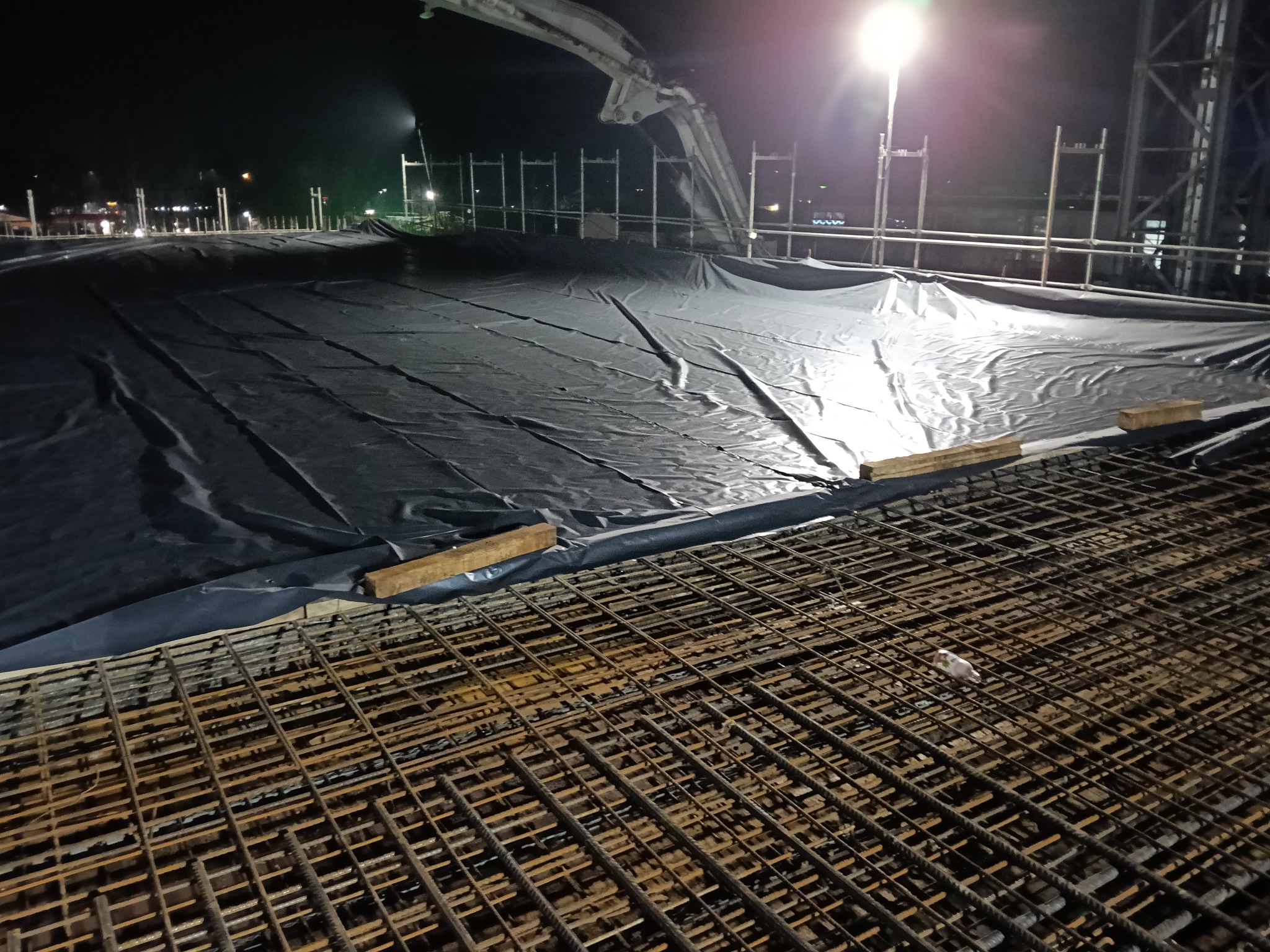 Nocny widok na zbrojenie fundamentów, zabezpieczone czarną folią. W tle widoczny dźwig do betonu i oświetlenie placu budowy. Detal zbrojenia na pierwszym planie.